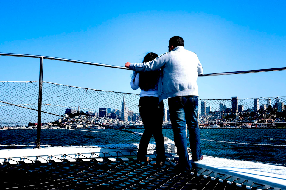 Two people standing on a boat's deck, looking at the city skyline across the water under a clear blue sky.