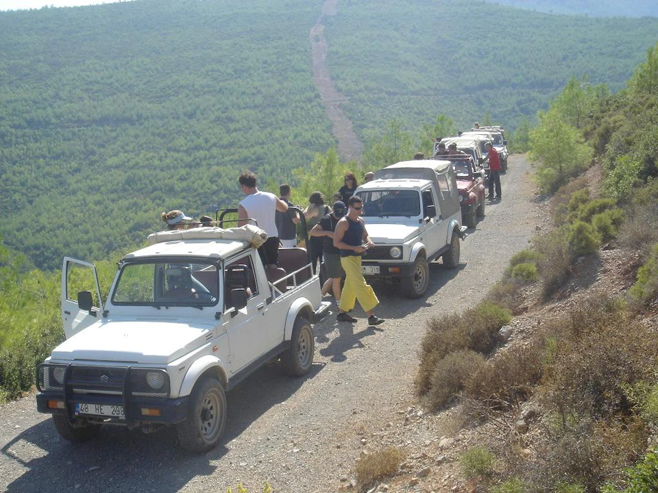 A group of people stands beside a line of off-road vehicles on a dirt road with a forested hill in the background.