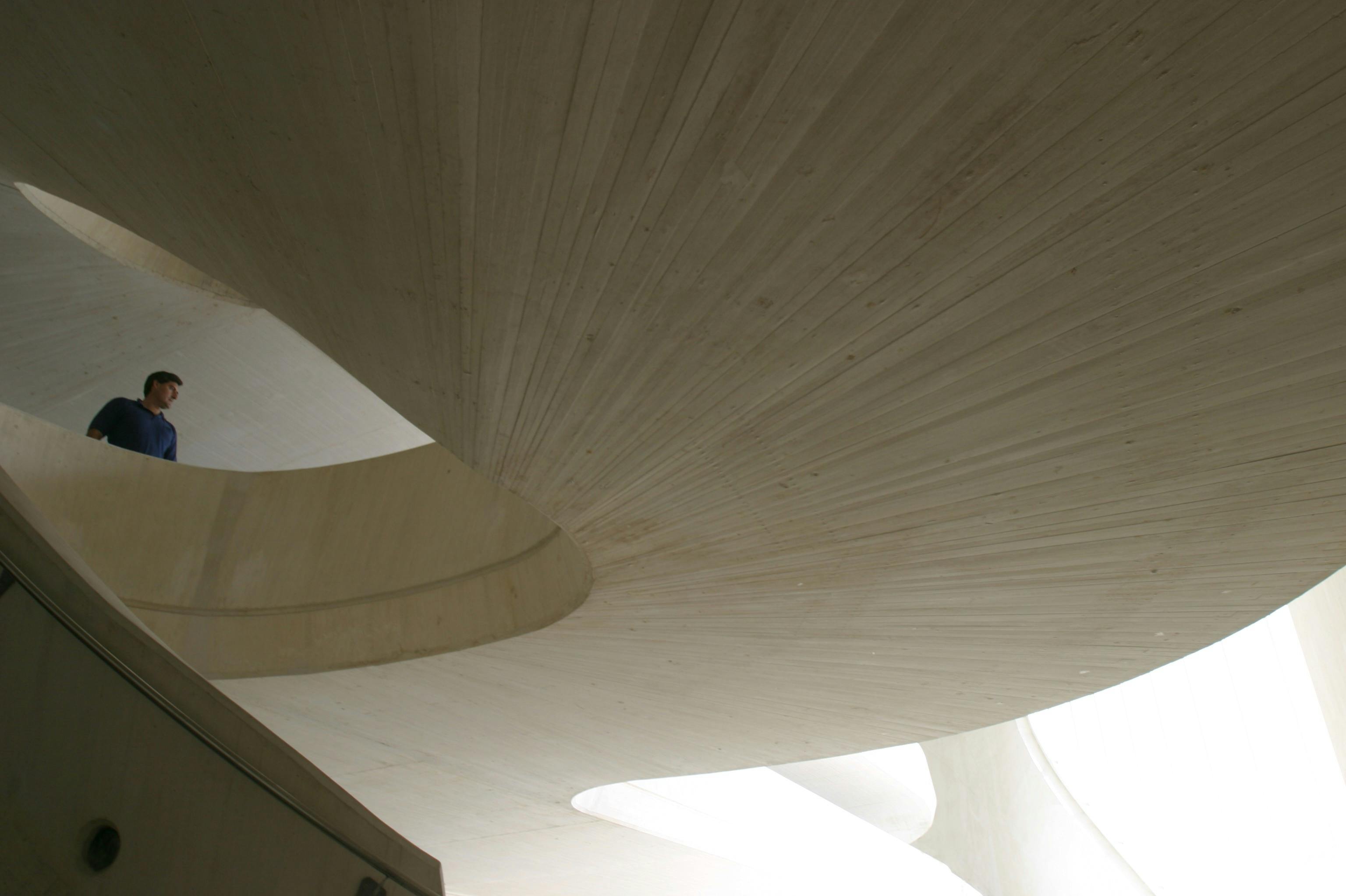 Zakřivená, světlá betonová konstrukce s moderním architektonickým designem, hladkými liniemi a protínajícími se rovinami.