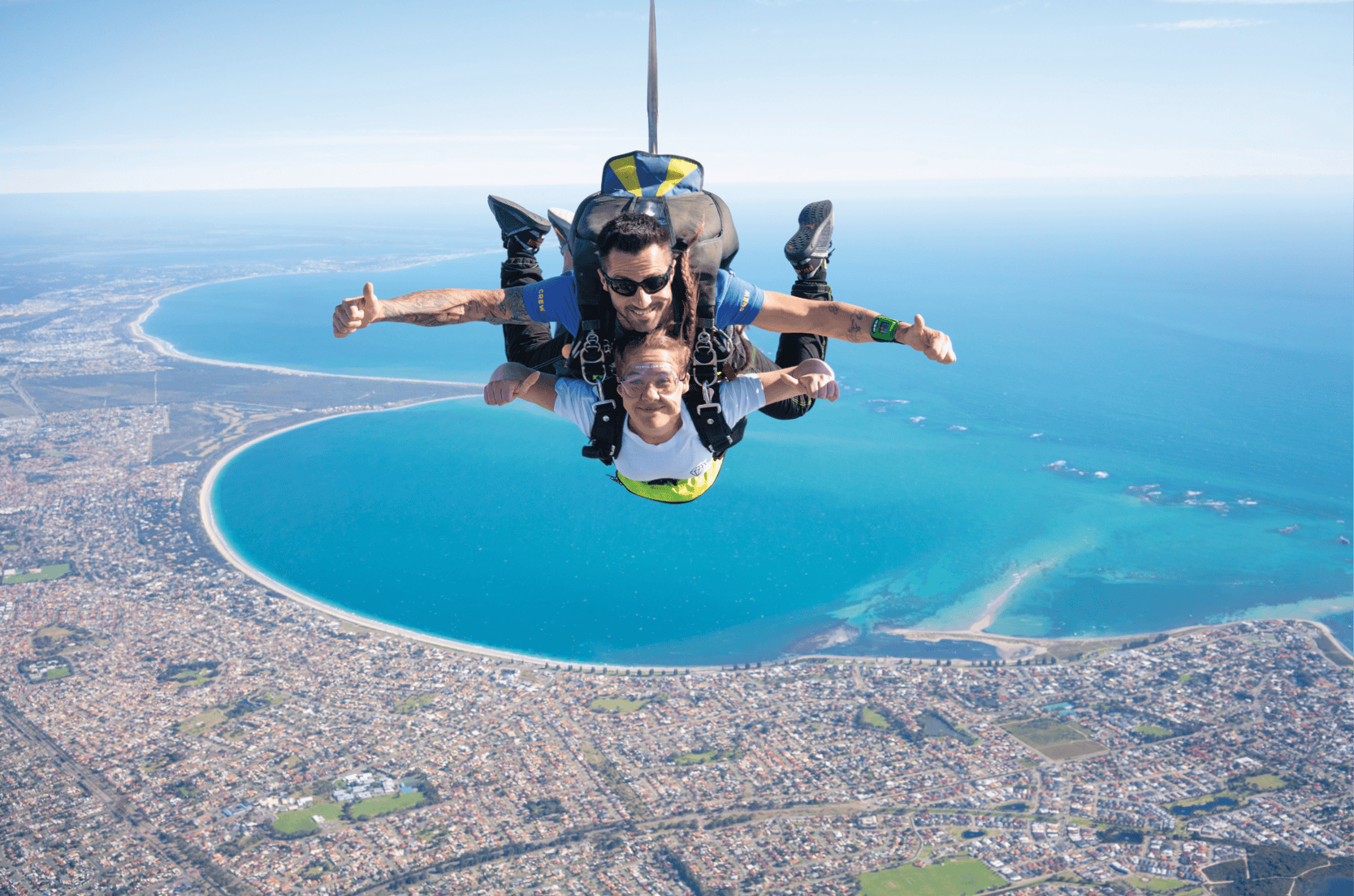 푸른 바다와 아래로 굽이진 해변이 보이는 해안 도시 위에서 두 사람이 탠덤 스카이다이빙을 즐기고 있습니다.