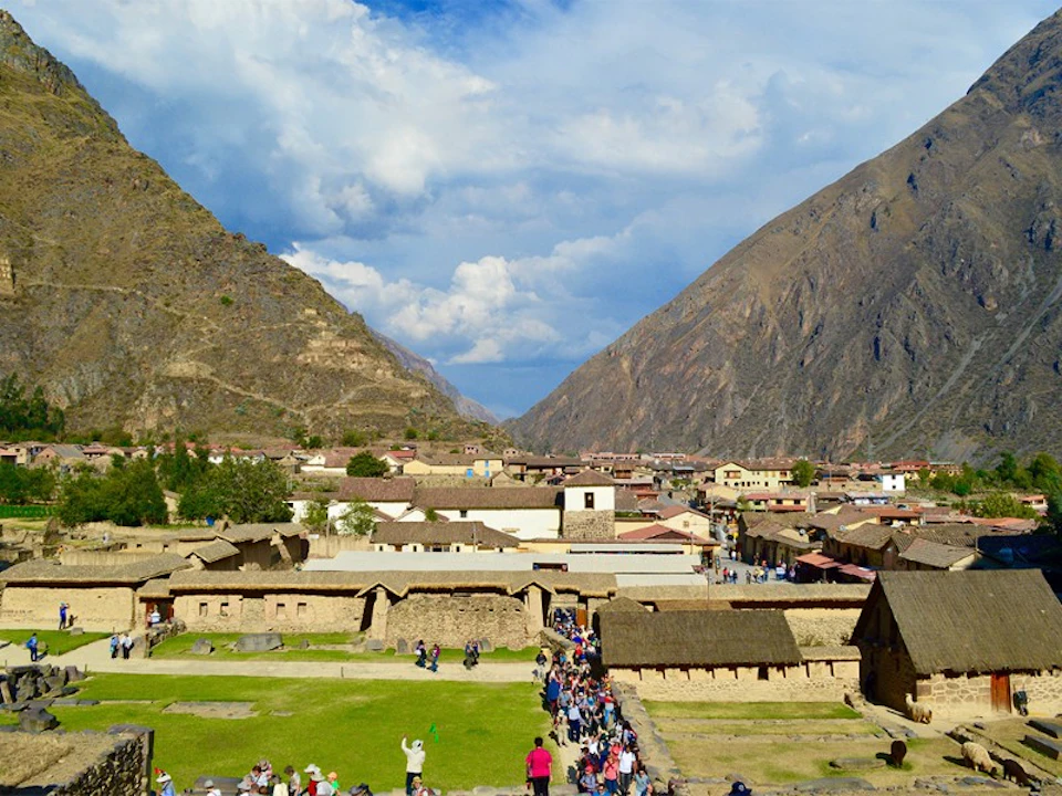 A scenic view of a village in a valley surrounded by mountains, with crowds of people walking through the pathways and stone buildings.