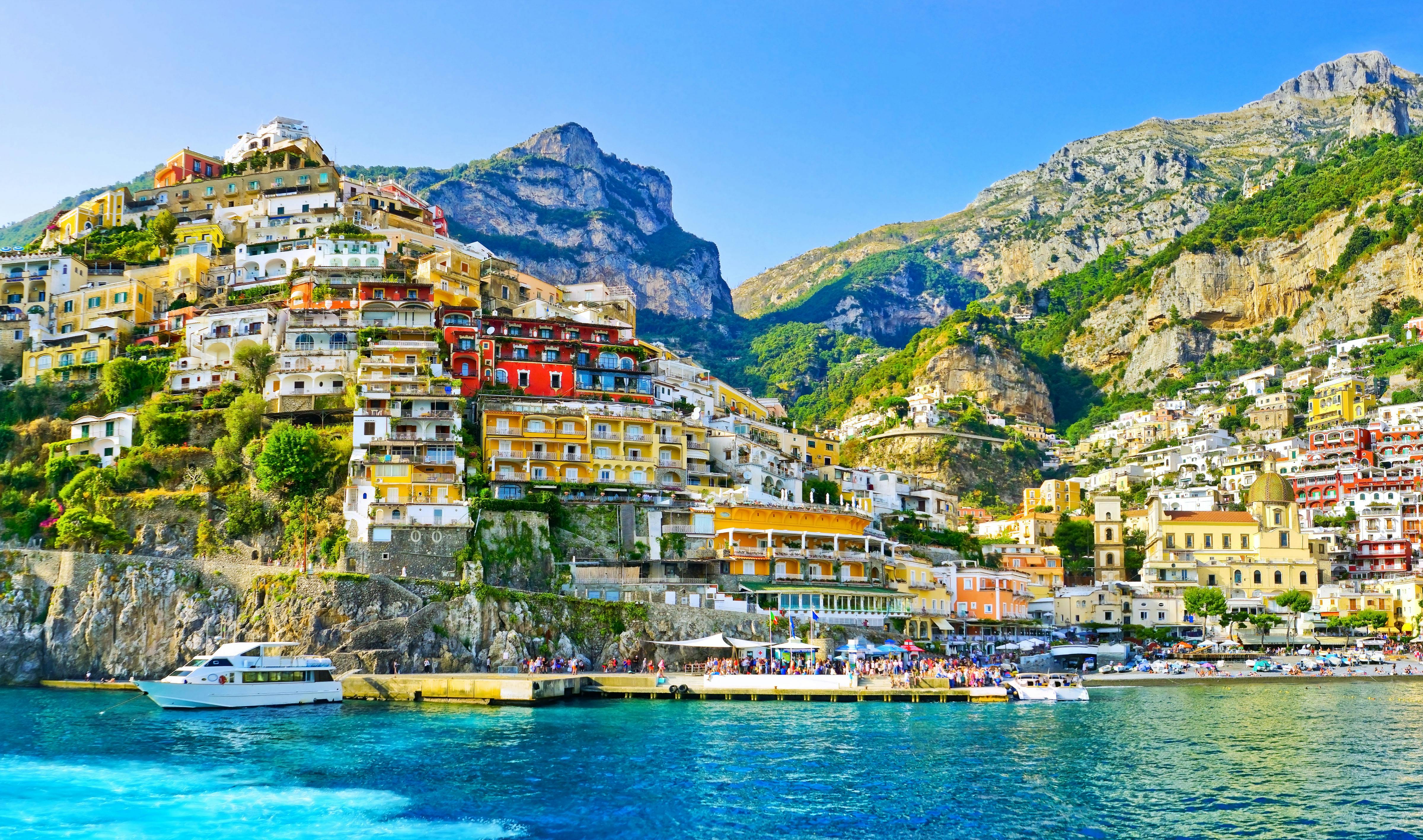 Colorful cliffside buildings in Positano, with boats docked along the waterfront and mountainous terrain in the background.