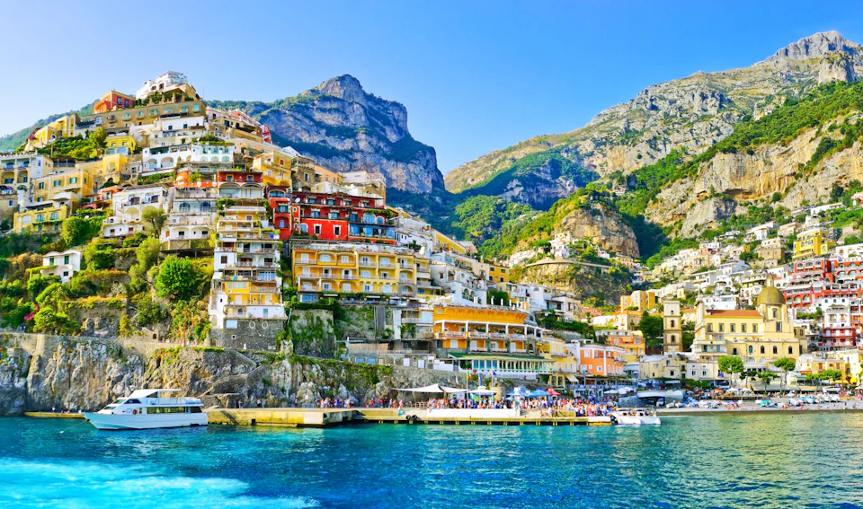 Colorful cliffside buildings in Positano, with boats docked along the waterfront and mountainous terrain in the background.