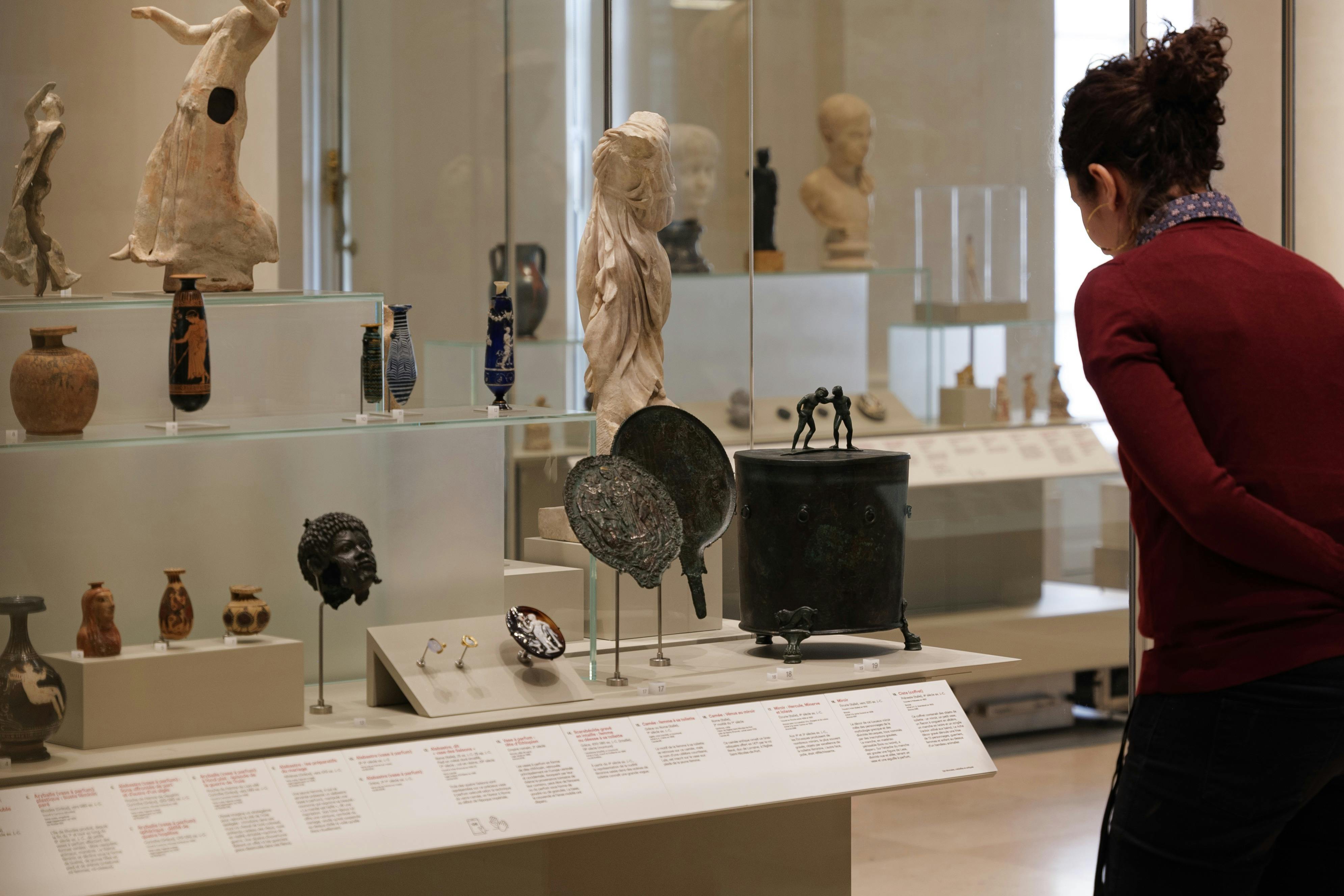 A person observing ancient artifacts displayed in a museum, including statues, pottery, and informational plaques.