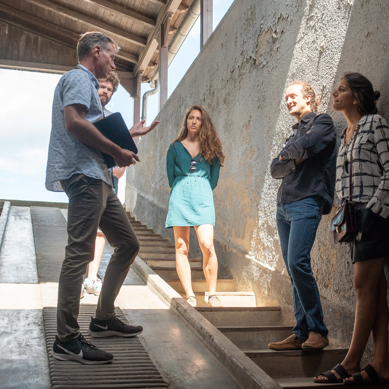 A group of people standing and talking near a concrete ramp in a sunlit, open structure.