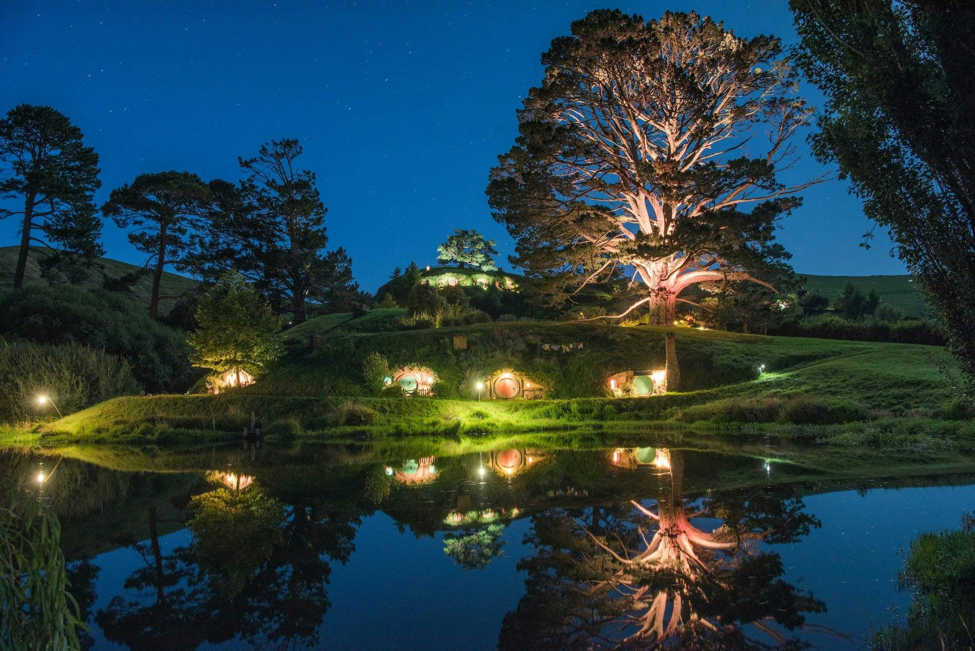 A serene nighttime scene with illuminated hobbit-style homes set into a grassy hill, a large lit tree, and a reflective pond.