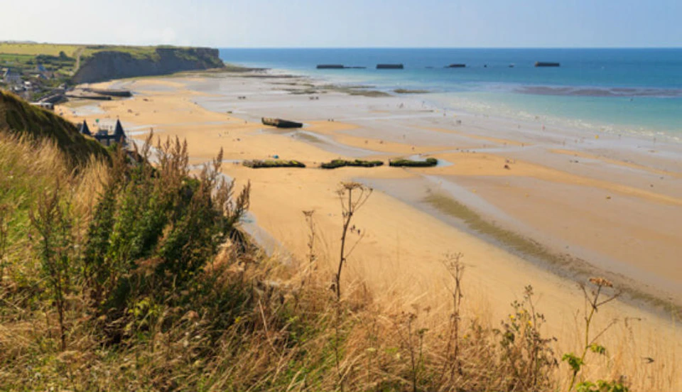 A sandy beach with scattered seaweed, people walking, and several concrete structures. Grass and shrubs in the foreground.