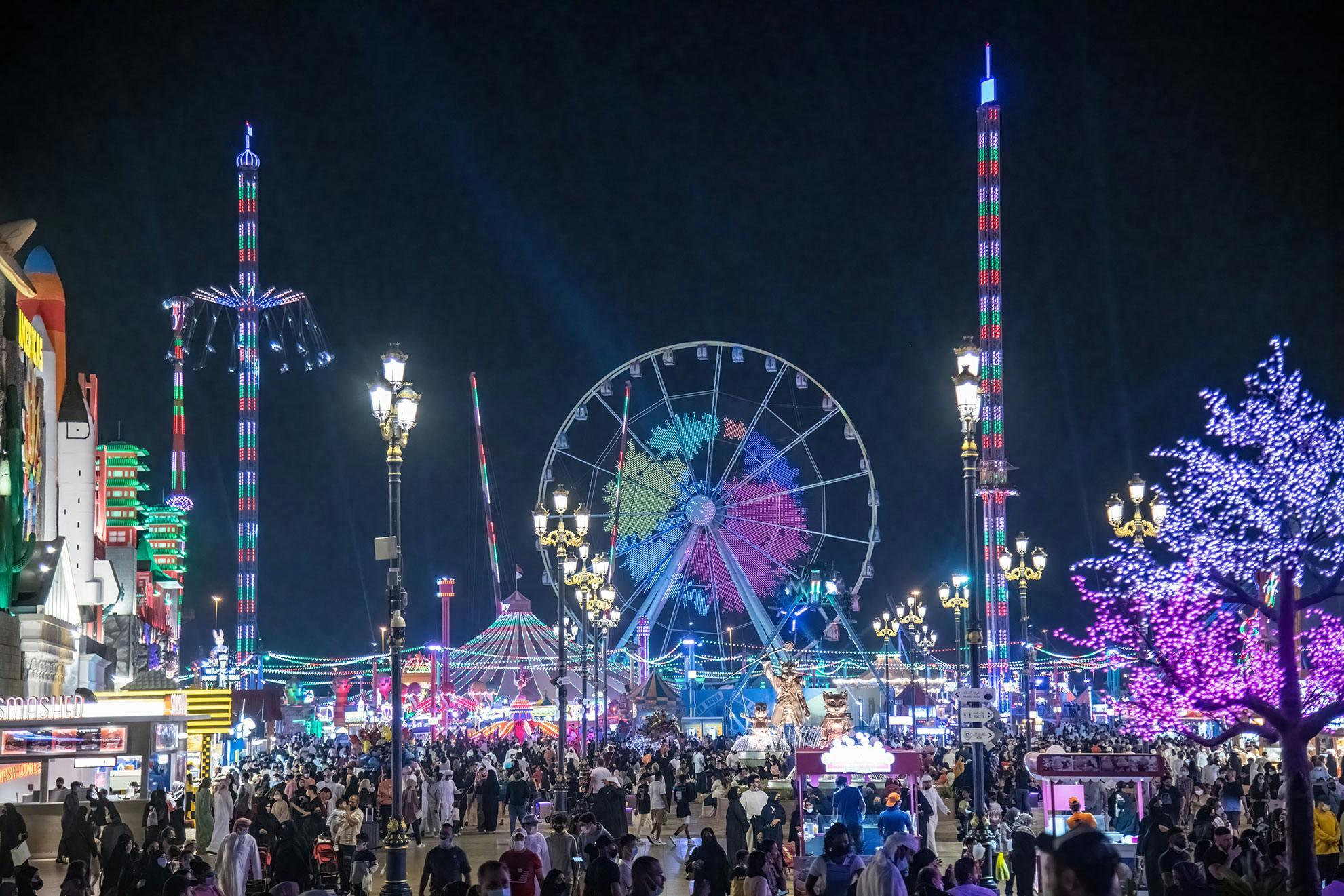 Rušný noční karneval s ruským kolem, vysokými jízdami, barevnými světly a velkým davem lidí, kteří si užívají slavnosti.