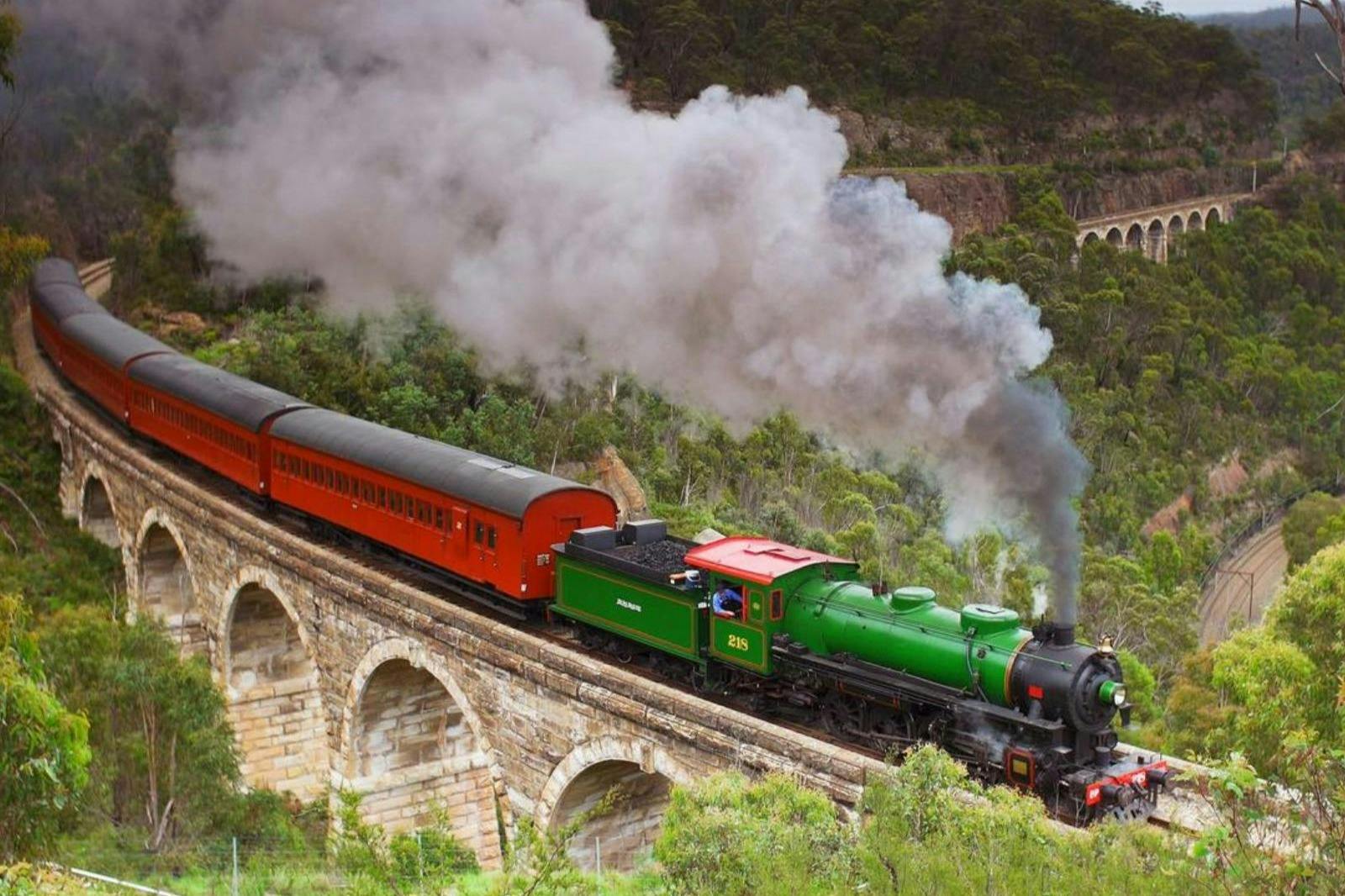 A green steam locomotive pulling red passenger cars crosses a stone bridge surrounded by lush greenery.