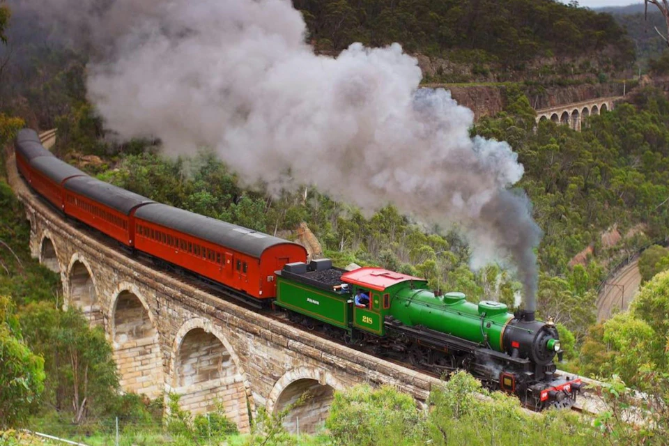 A green steam locomotive pulling red passenger cars crosses a stone bridge surrounded by lush greenery.