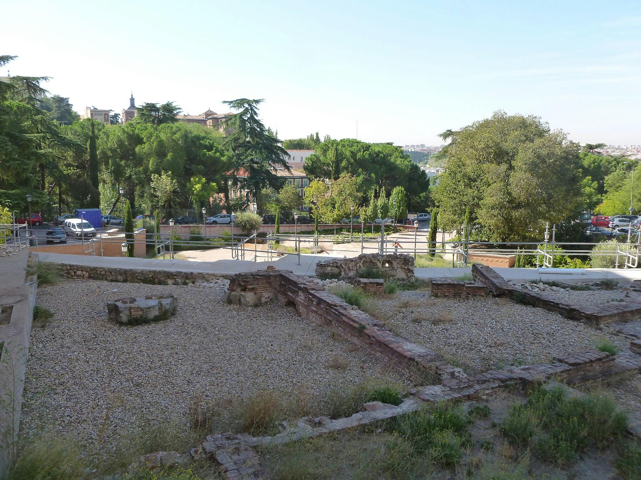 Starobylé kamenné zříceniny obklopené zábradlím, svěžími stromy a zaparkovanými vozidly v městském parku pod jasnou oblohou.