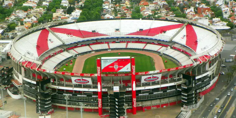 Vue aérienne d'un grand stade vide avec des sièges rouges et blancs, entouré de bâtiments urbains et de verdure.
