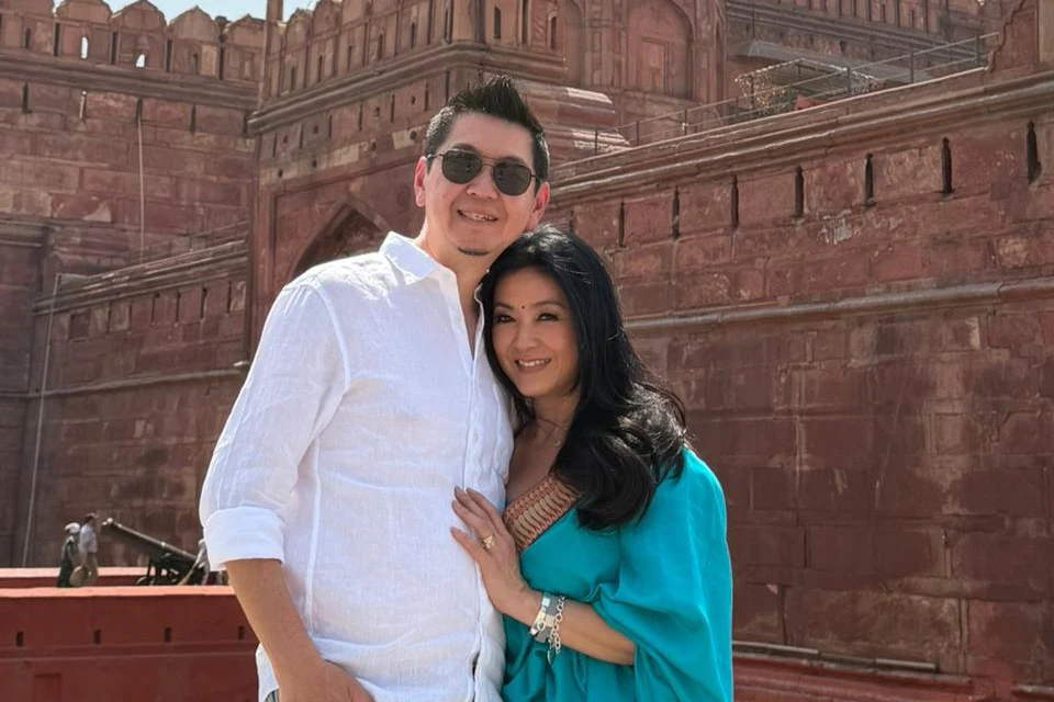 A man and a woman posing together in front of a red stone historical building under bright sunlight.