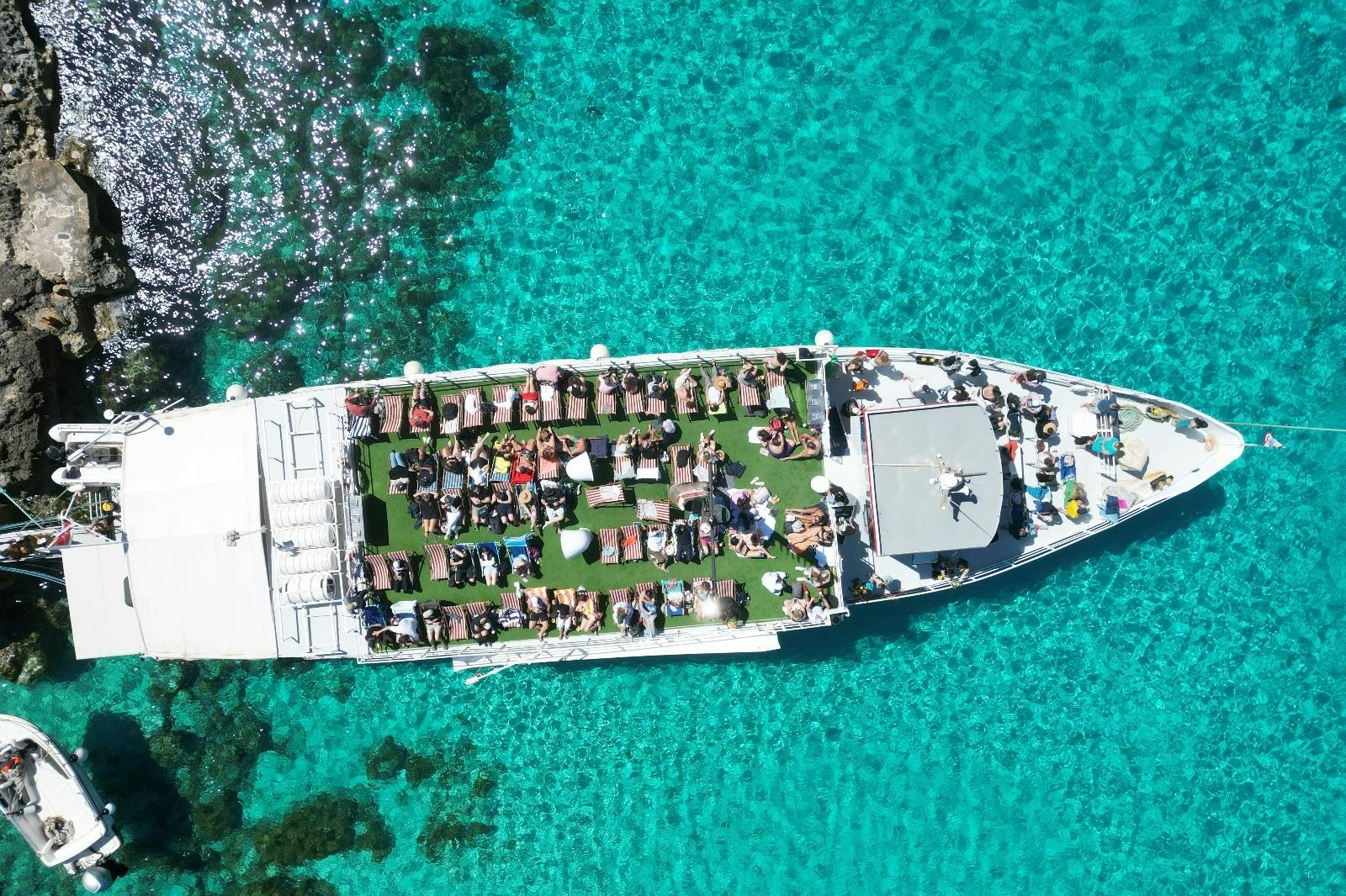Εναέρια άποψη ενός σκάφους σε καθαρά γαλαζοπράσινα νερά, με τους ανθρώπους να κάνουν ηλιοθεραπεία στις ξαπλώστρες στο επάνω κατάστρωμα.