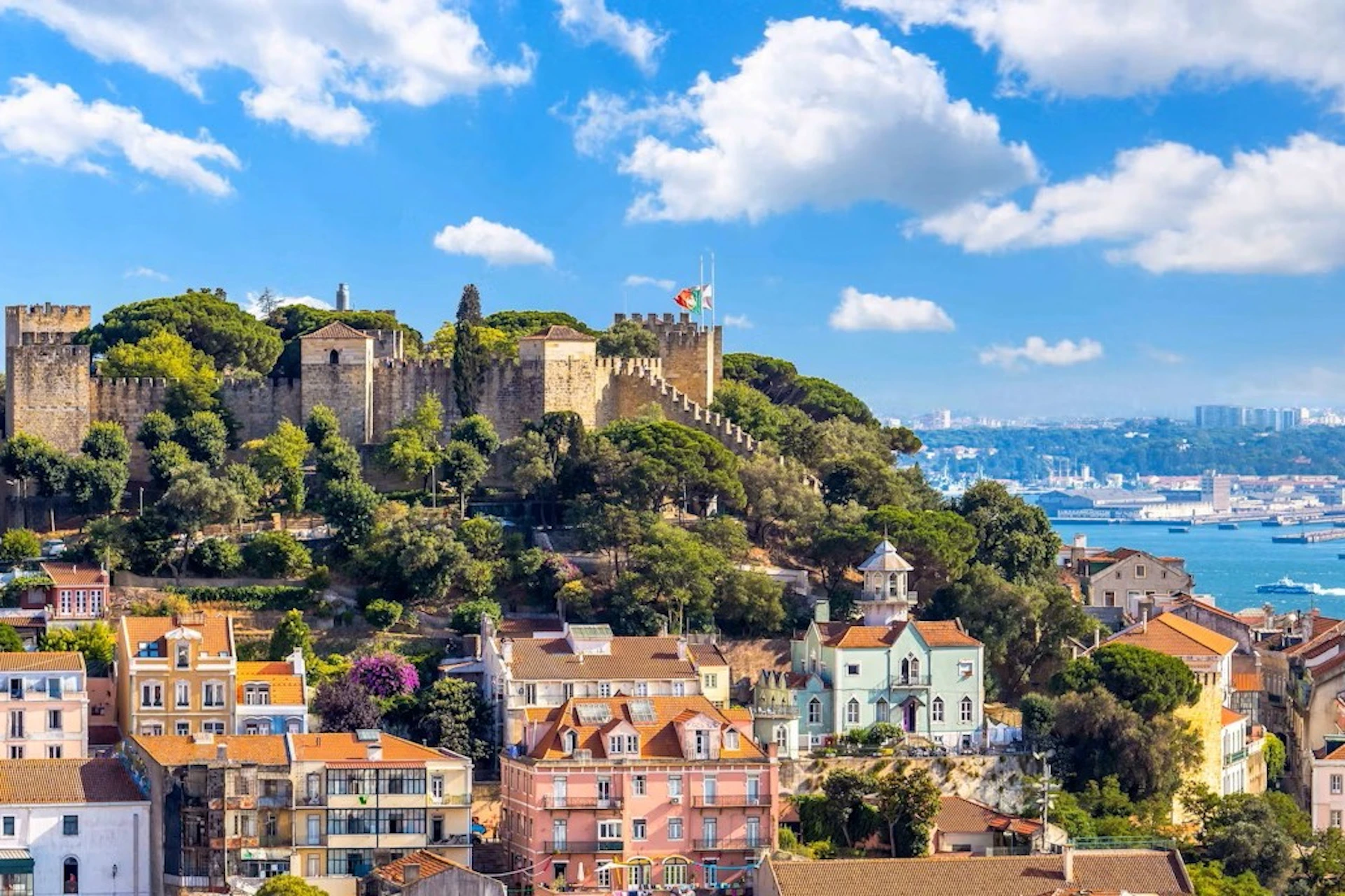 A castle on a hill surrounded by trees and colorful houses under a blue sky with scattered clouds.