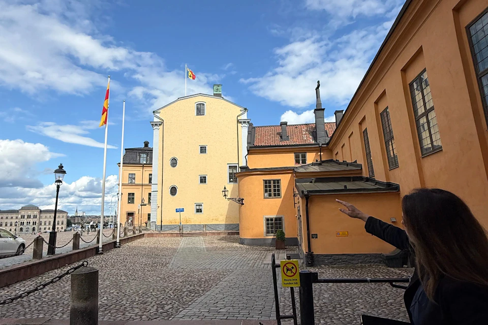 Courtyard with cobblestone ground, yellow buildings, flags, and a person gesturing on the right side under a blue sky.