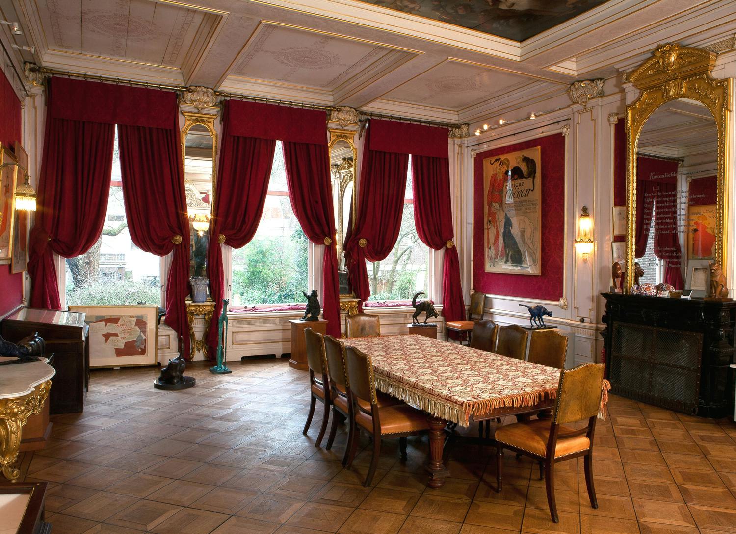 房间布置优雅，配有长餐桌、华丽的椅子、红色窗帘、大窗户和墙上的框架艺术品。
