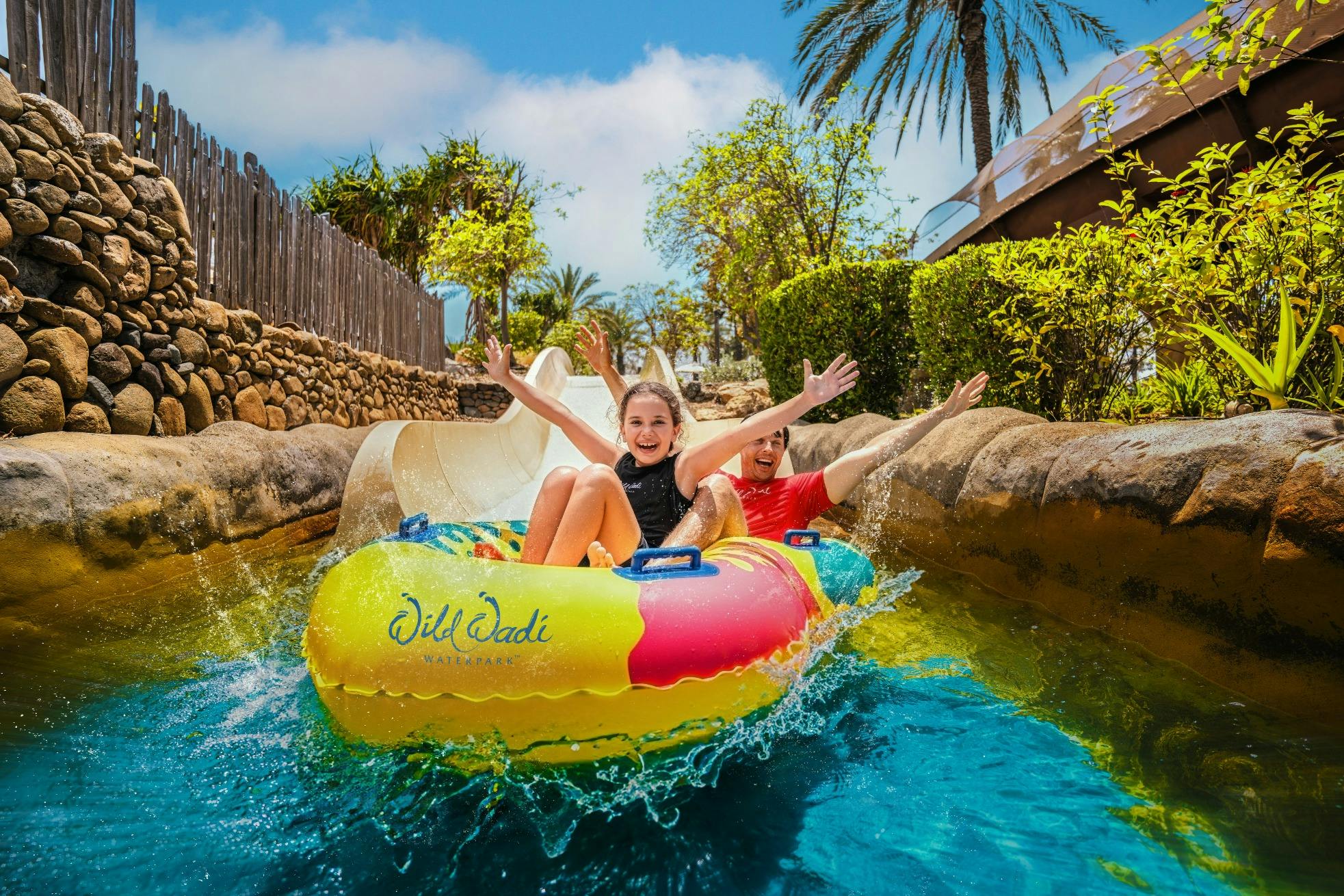 Deux personnes sur un radeau gonflable coloré glissant sur un manège aquatique dans un parc en plein air, entouré de plantes et de palmiers.