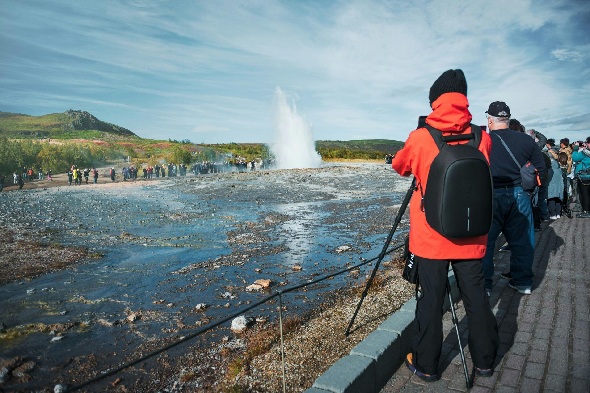 A person in an orange jacket with a camera on a tripod photographing a geyser erupting, with onlookers in the background.