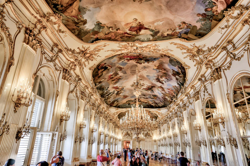 A grand hall with ornate chandeliers, detailed ceiling frescoes, and visitors observing the decor.