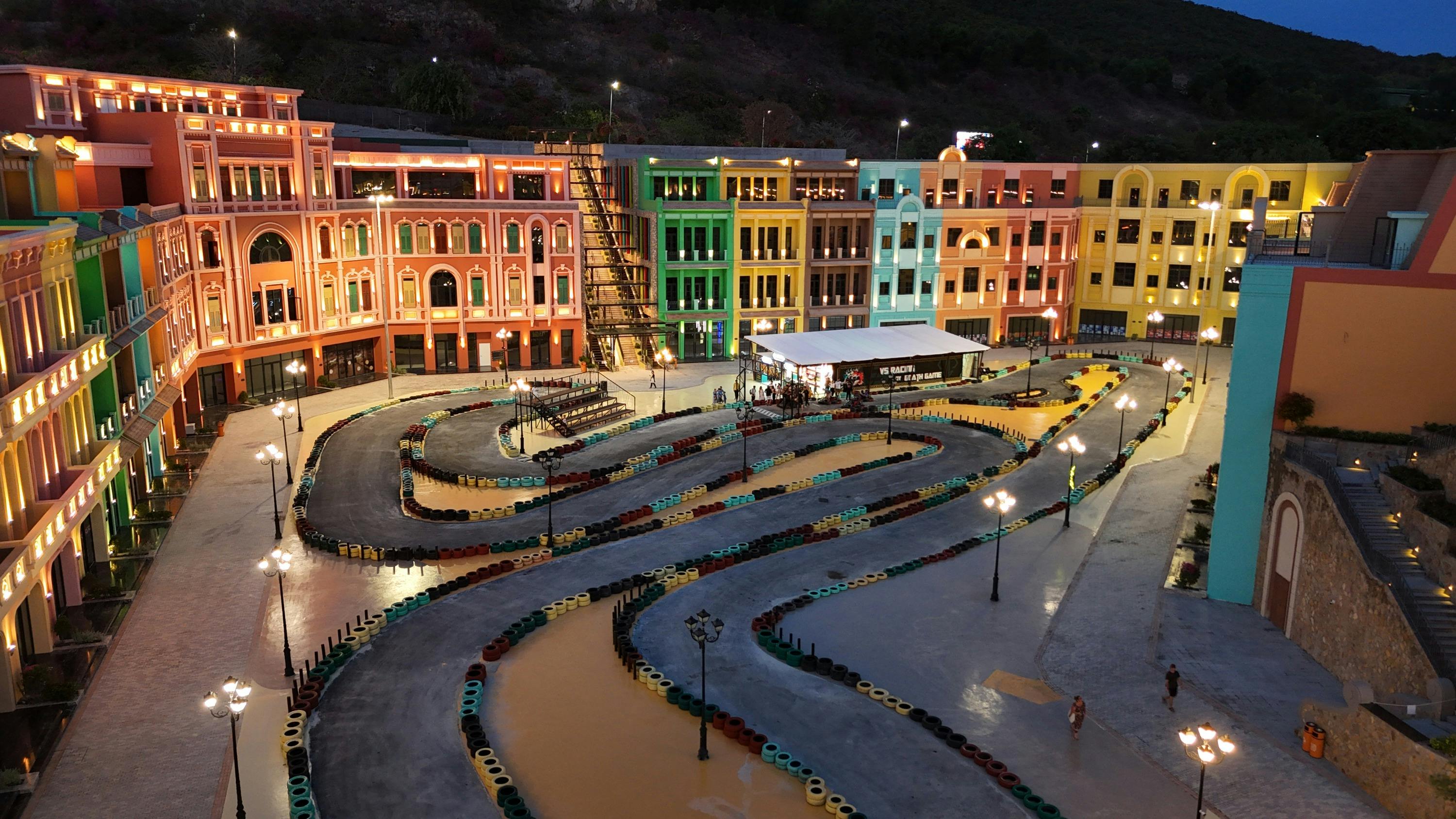 Une piste de karting illuminée, entourée de bâtiments colorés et animés à la tombée de la nuit, avec quelques personnes qui se promènent à proximité.