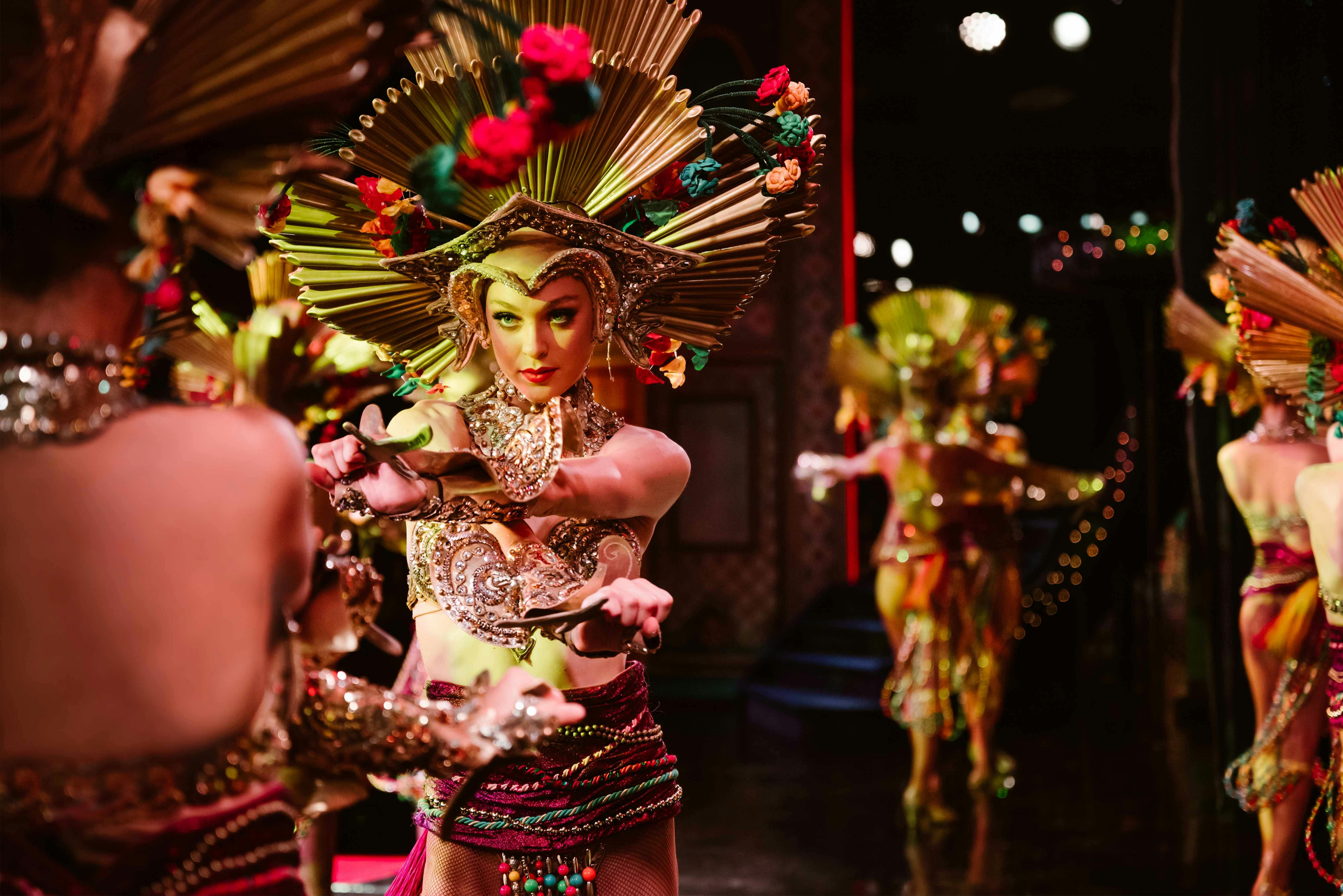 A performer in colorful, ornate attire and a headdress in a theatrical setting, extending her arms in a dance pose.