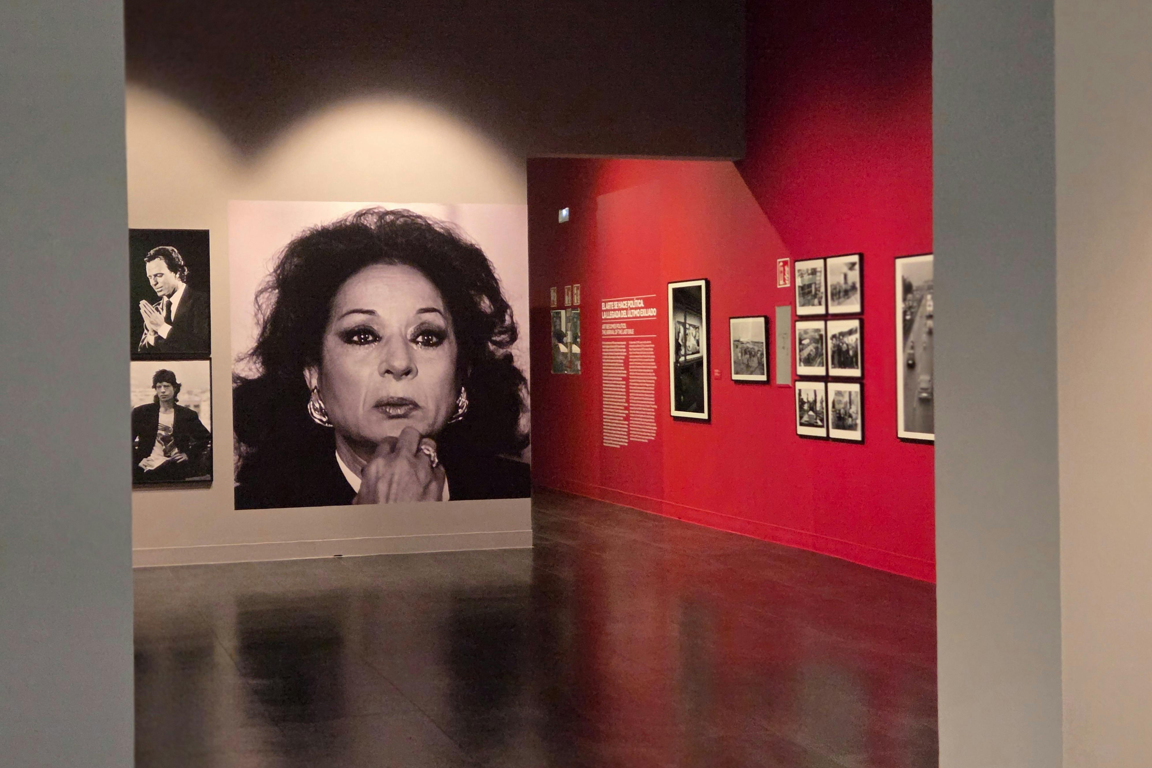 Uma exposição de museu com um grande retrato a preto e branco de uma mulher e fotografias mais pequenas em paredes adjacentes com detalhes em vermelho.