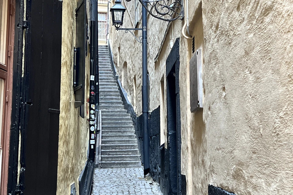 Narrow, cobblestone alley with steep stairs leading up between old, textured buildings, and adorned with wall lamps.