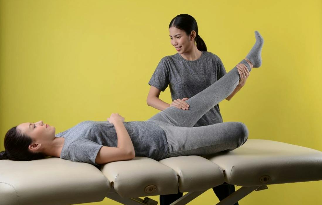 A therapist is stretching a person's leg on a massage table against a yellow background.