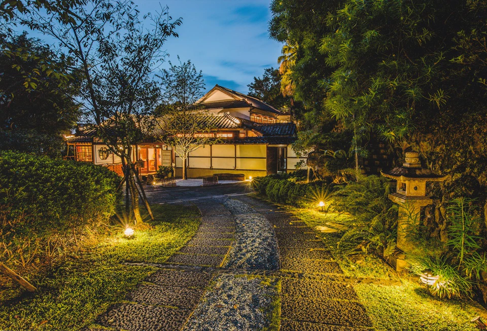 A traditional Japanese house with lit garden pathway under a twilight sky, surrounded by trees and lanterns.
