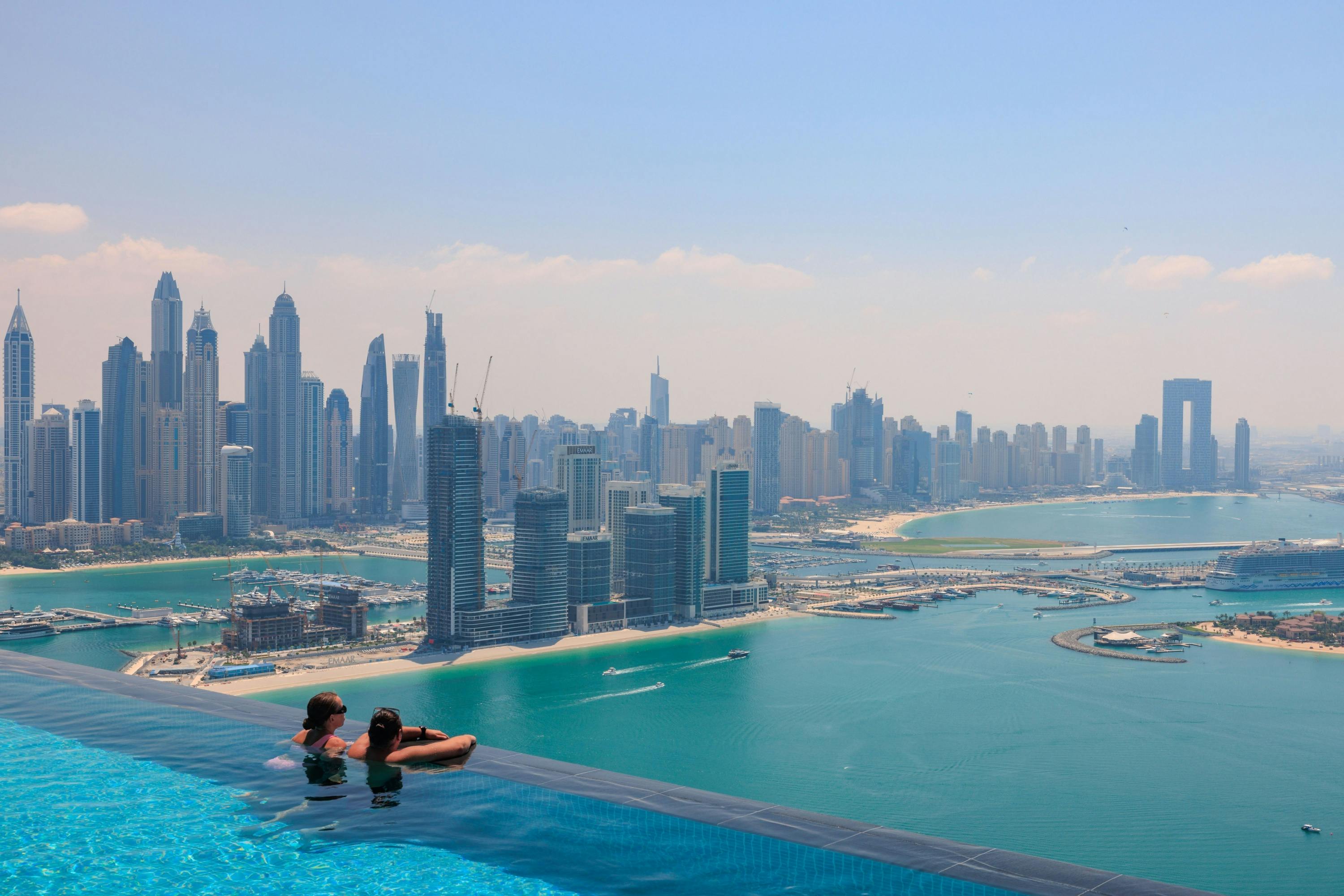 Due persone si rilassano in una piscina a sfioro che si affaccia su una baia e sullo skyline di una città moderna con numerosi edifici alti.