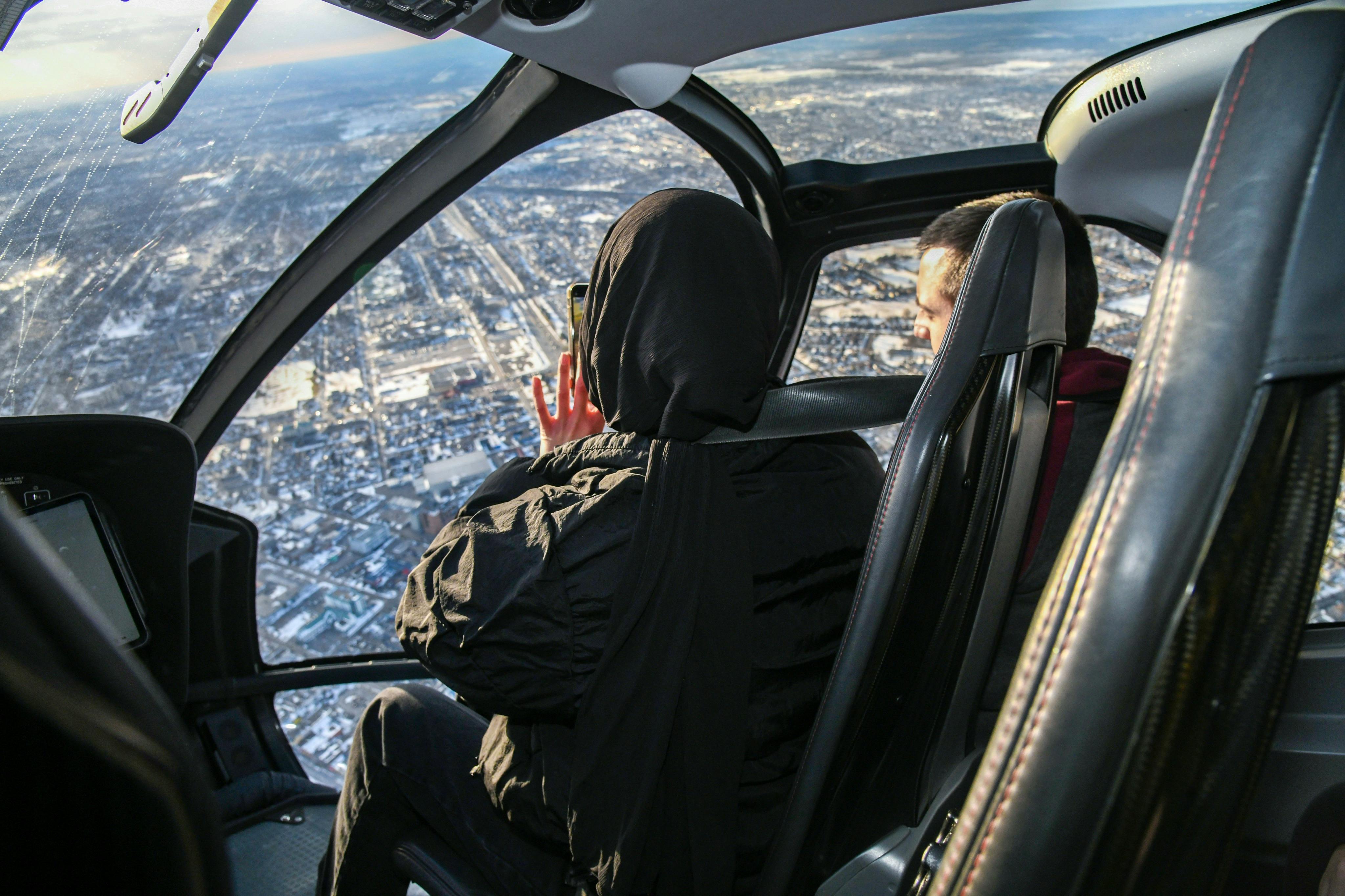 Two people inside a helicopter, one taking photos of the snowy cityscape below through the window.