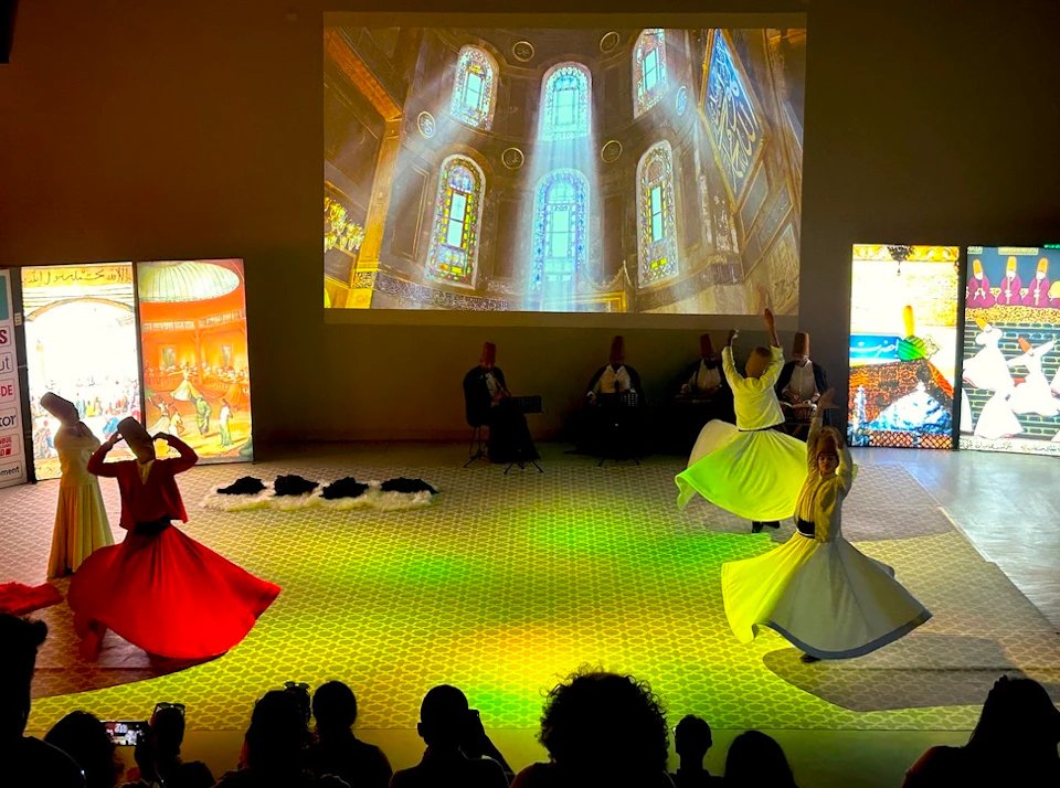 People in traditional attire perform a Sufi dance on a patterned floor, with a large projection of stained glass windows in the background.