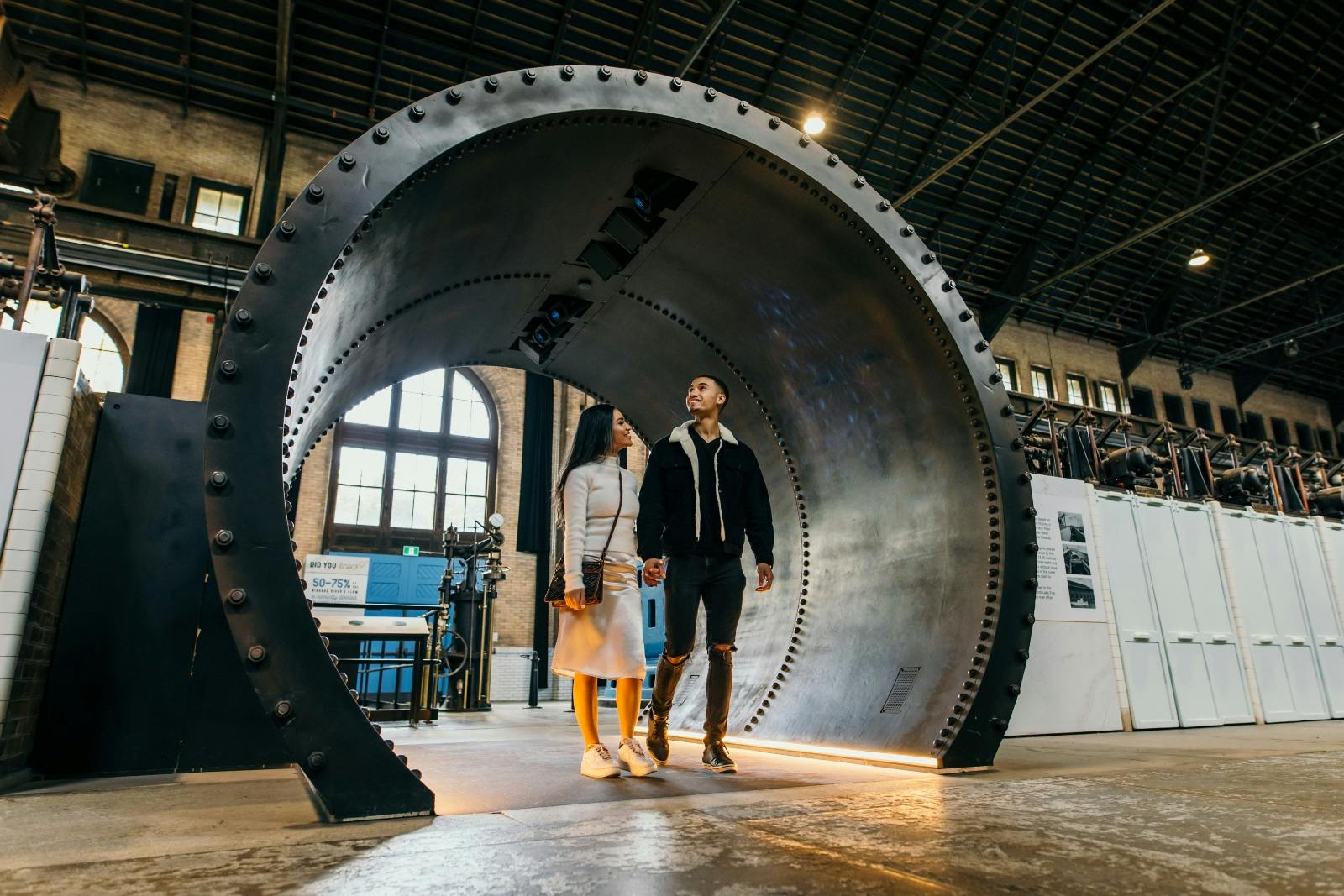 To personer, der går gennem en stor cirkulær stålkonstruktion inde i en bygning i industriel stil med højt til loftet og store vinduer.