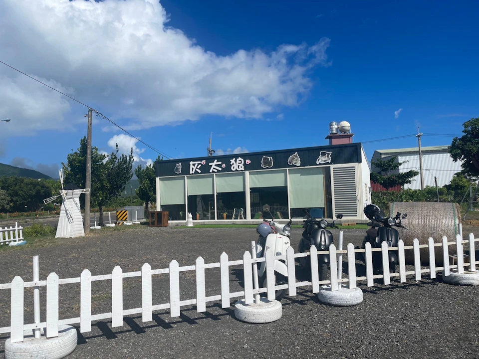 A sunny day view of a modern building with large glass windows, signage, a white fence, and scooters parked outside.