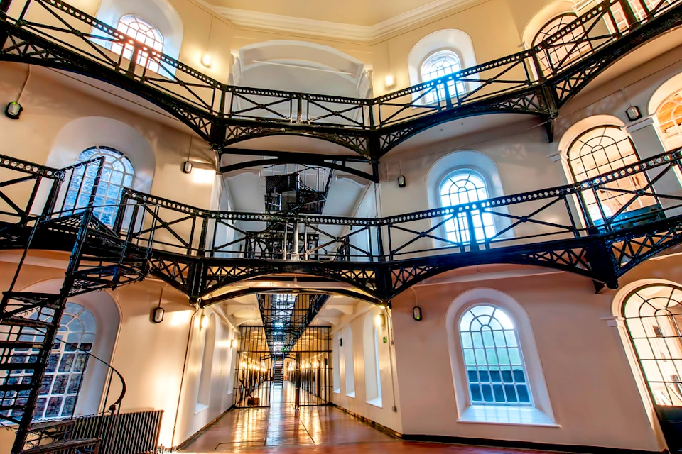 A multi-level interior with metal walkways, arched windows, and decorative lighting, resembling an old prison or institutional building.