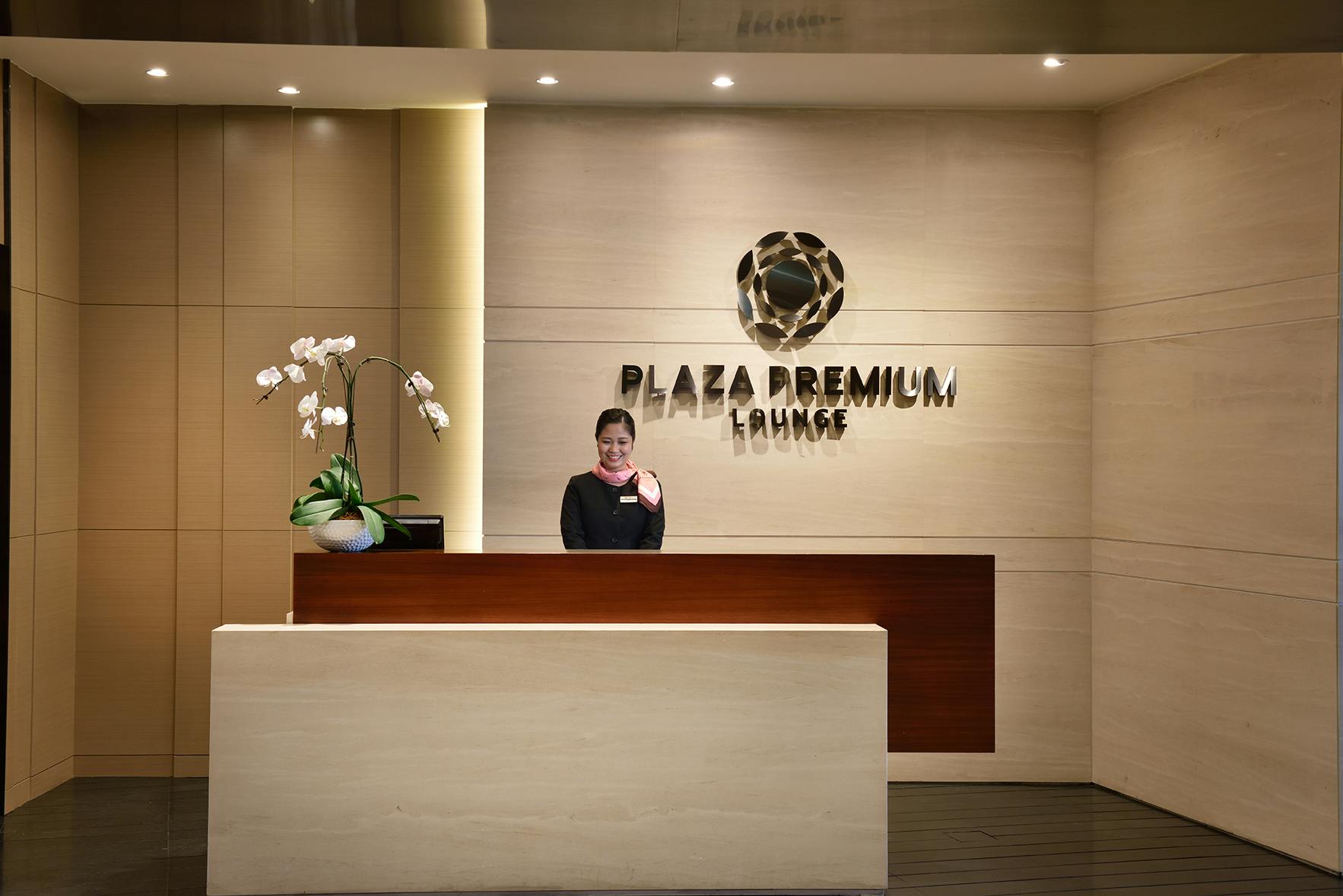 Un taulell de recepció amb un amable personal, planta d'orquídies i rètol de "Plaza Premium Lounge" a la paret de darrere.