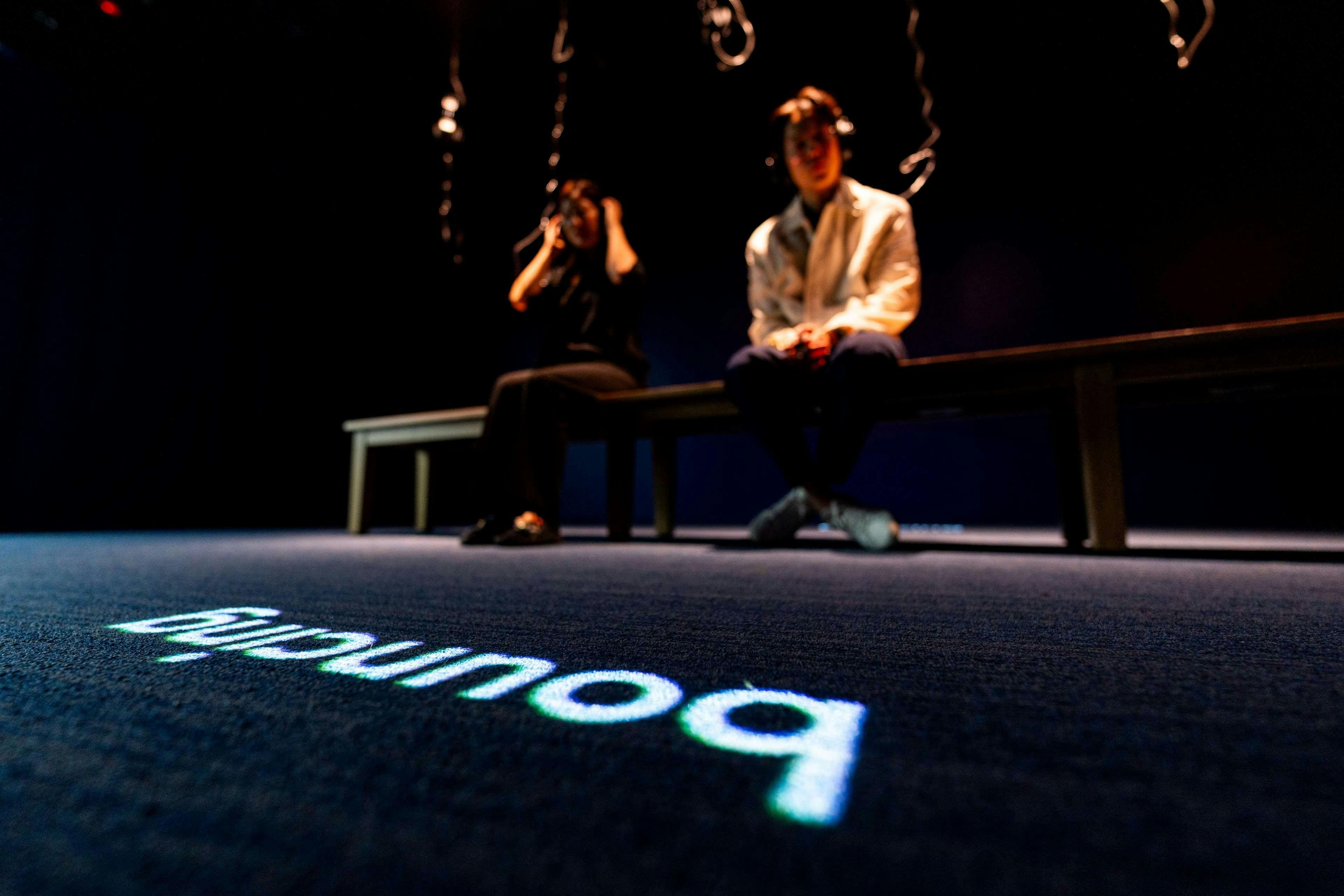Two people sitting on a bench in a dark room, wearing headphones, with the word "boundary" projected on the floor.