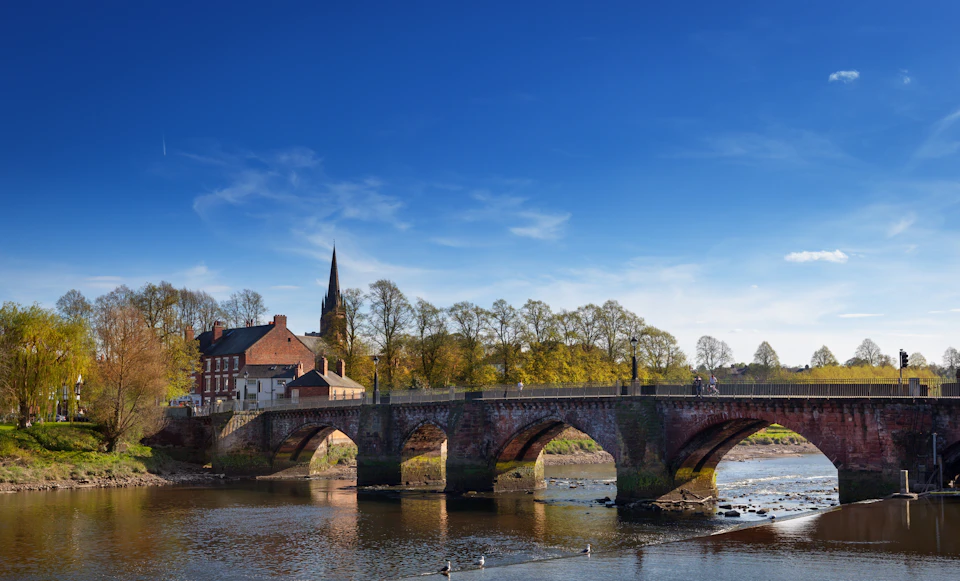 A stone bridge with arches over a river, with buildings and a church spire in the background under a clear blue sky.