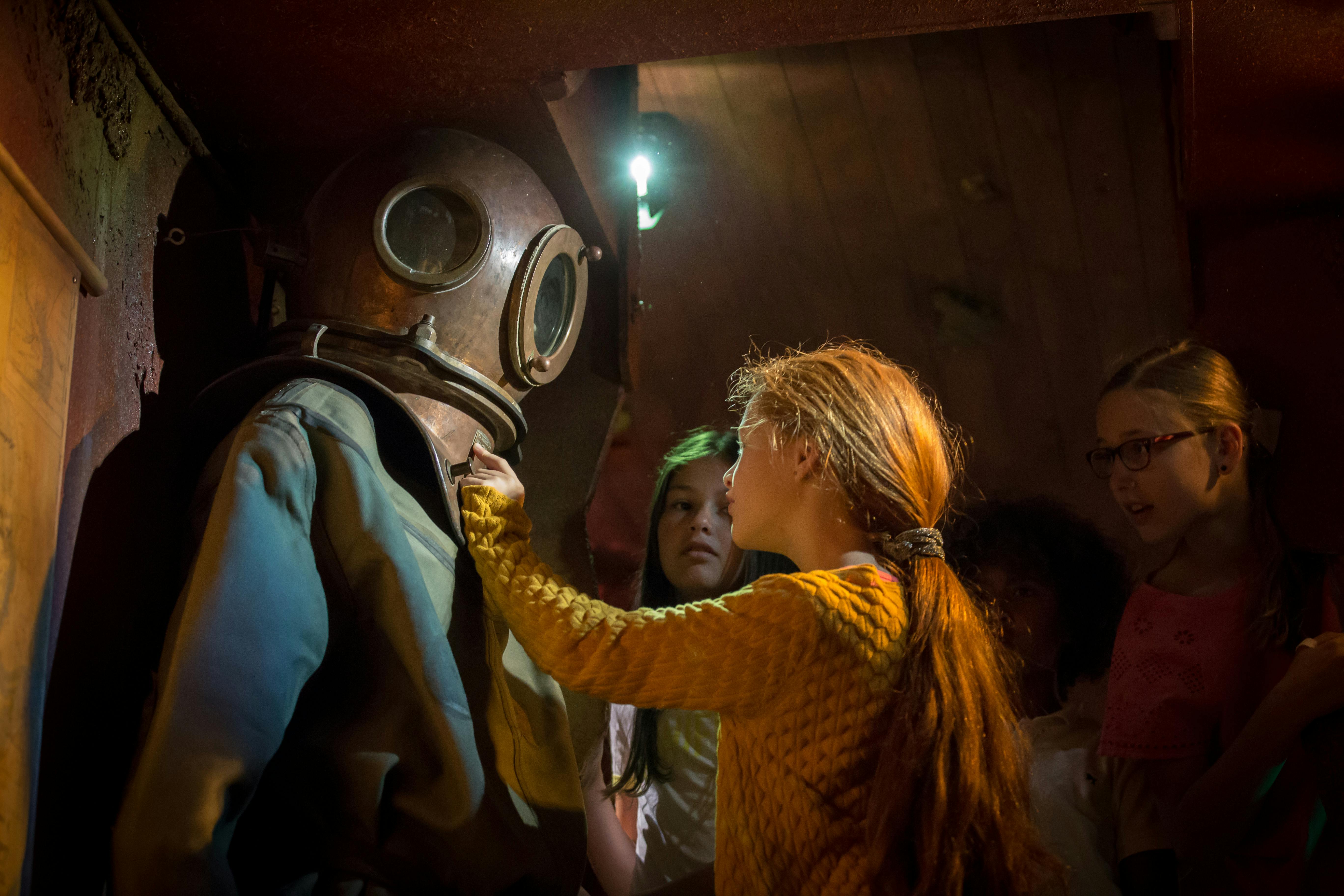 Skupina dětí, včetně dívky s rudými vlasy, zkoumá staromódní potápěčský oblek a helmu v tlumeně osvětleném prostředí.