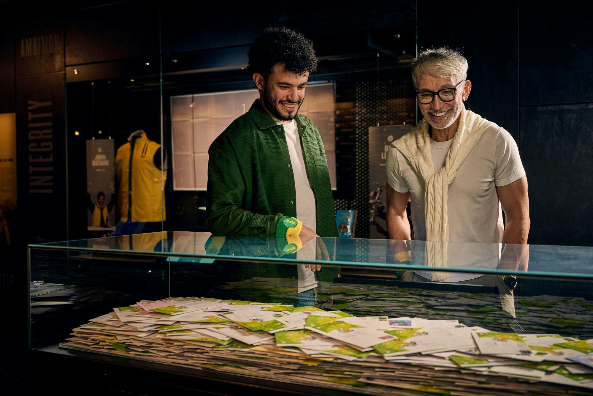 Deux hommes sourient devant une vitrine contenant divers objets dans un décor intérieur moderne.