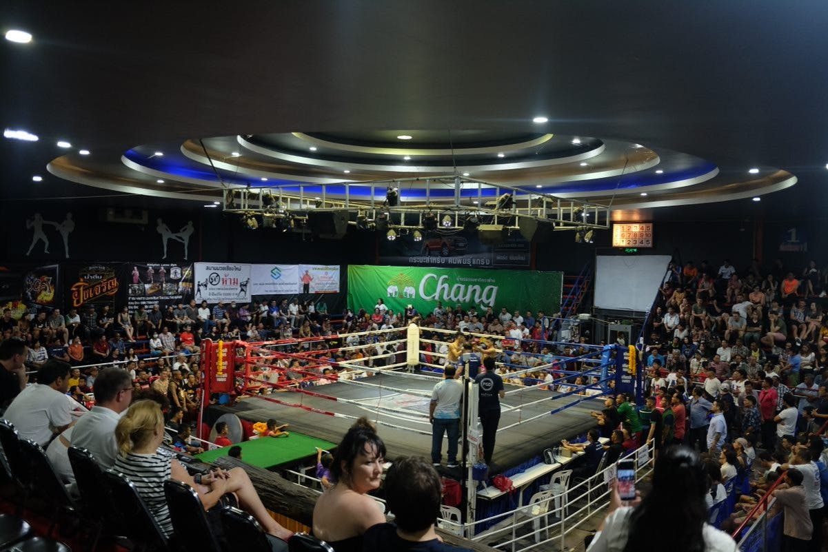 Une arène intérieure bondée avec un ring de boxe au centre, entouré de spectateurs et de bannières sur les murs.