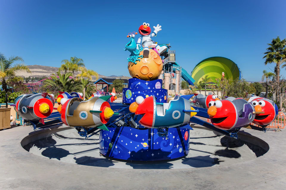 Elmo-themed amusement park ride with rocket ships on rotating arms. Palm trees, slides, and a dome structure in the background.