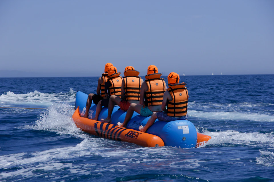 Four people in life jackets riding an inflatable banana boat on the ocean; clear blue sky in the background.