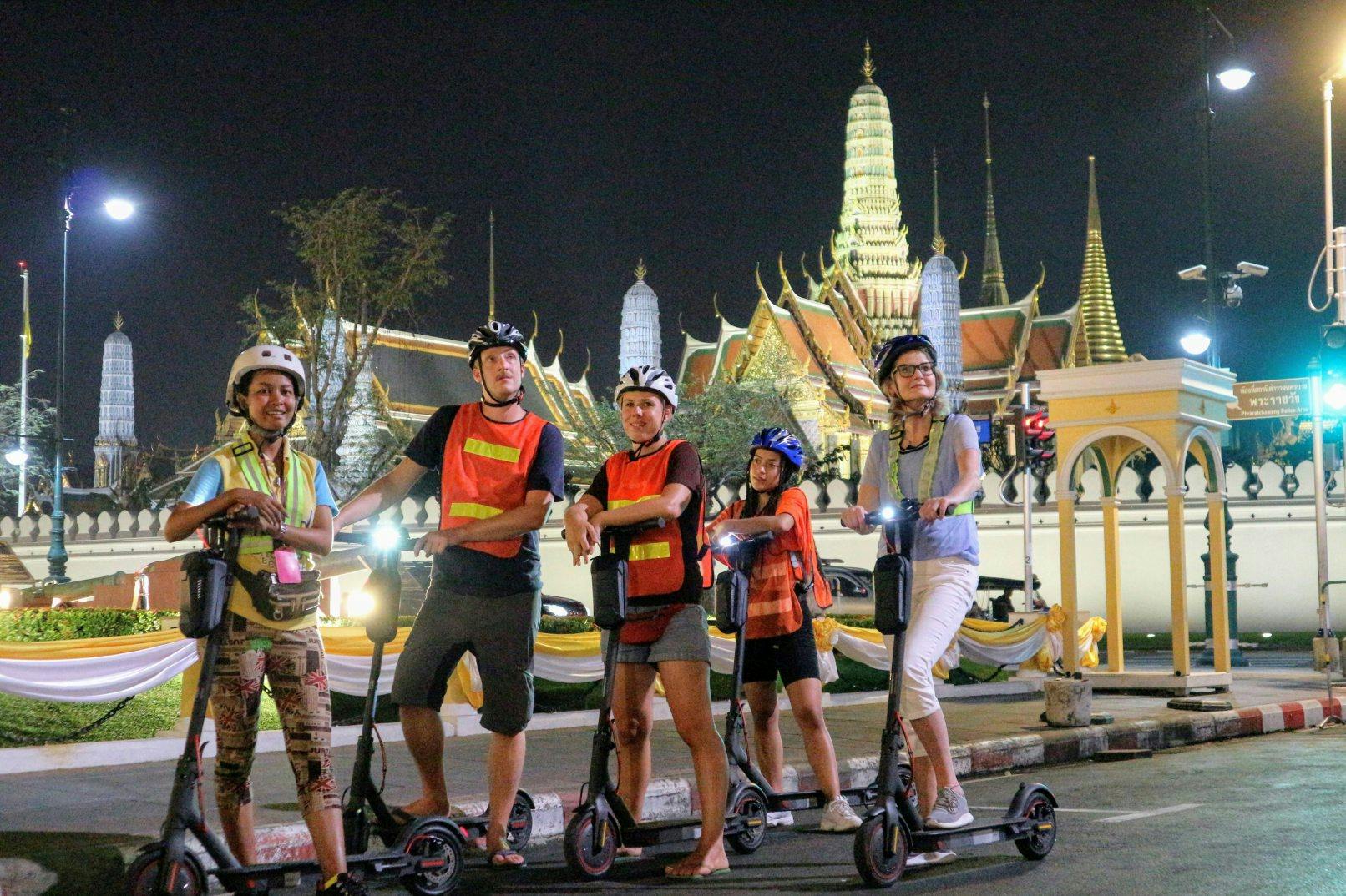 A group of five people on electric scooters in front of a lit temple at night.