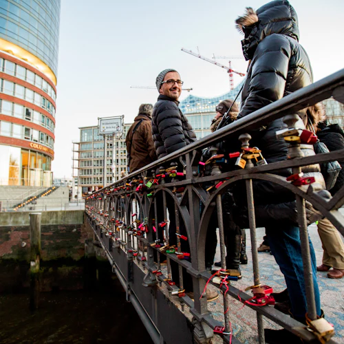 Människor som står på en bro prydd med kärlekslås, med byggnader och byggkranar synliga i bakgrunden.