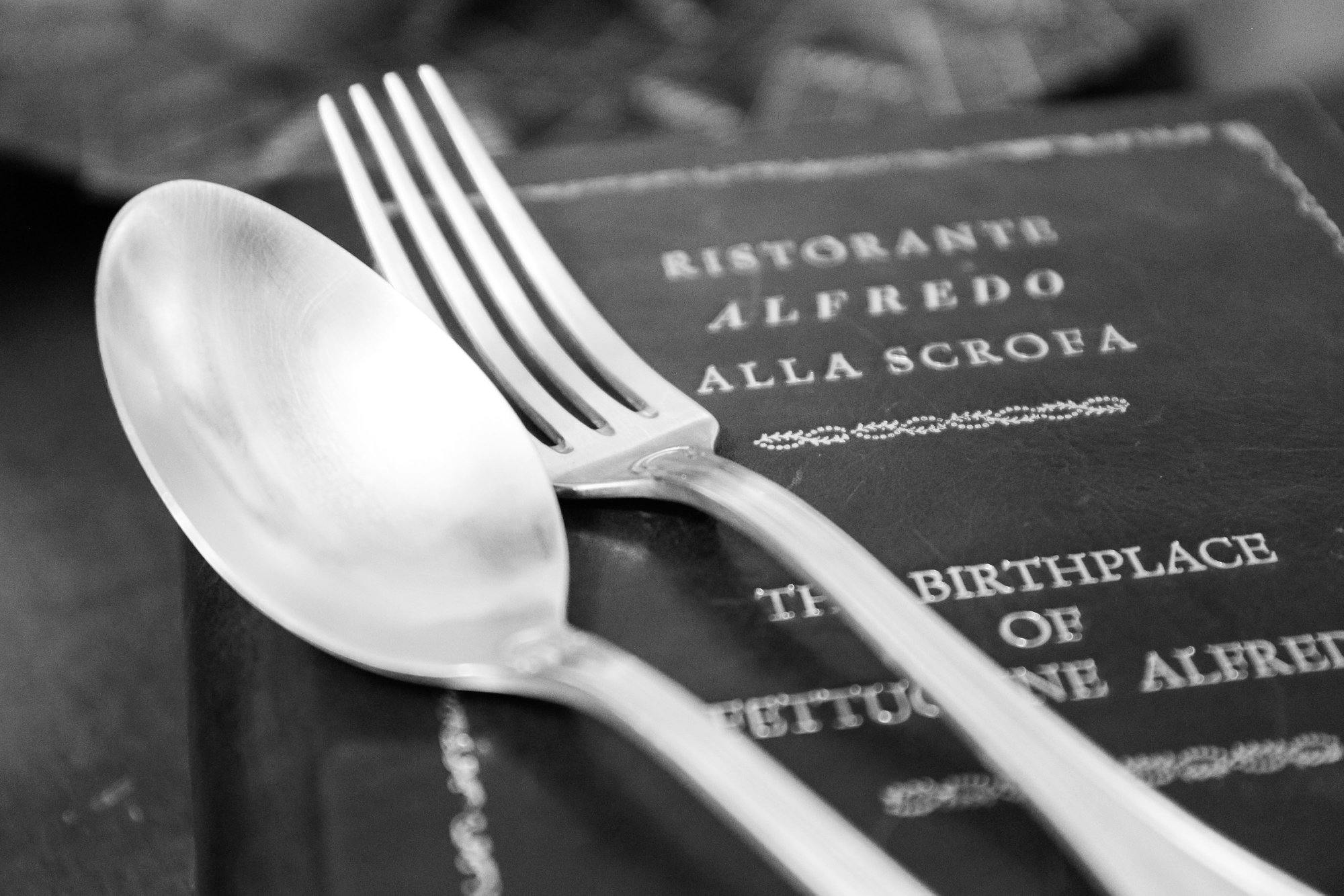 Крупный план ложки и вилки, опирающихся на книгу с видимым текстом: «RISTORANTE ALFREDO ALLA SCROFA» и «THE BIRTH OF FETTUCCINE».