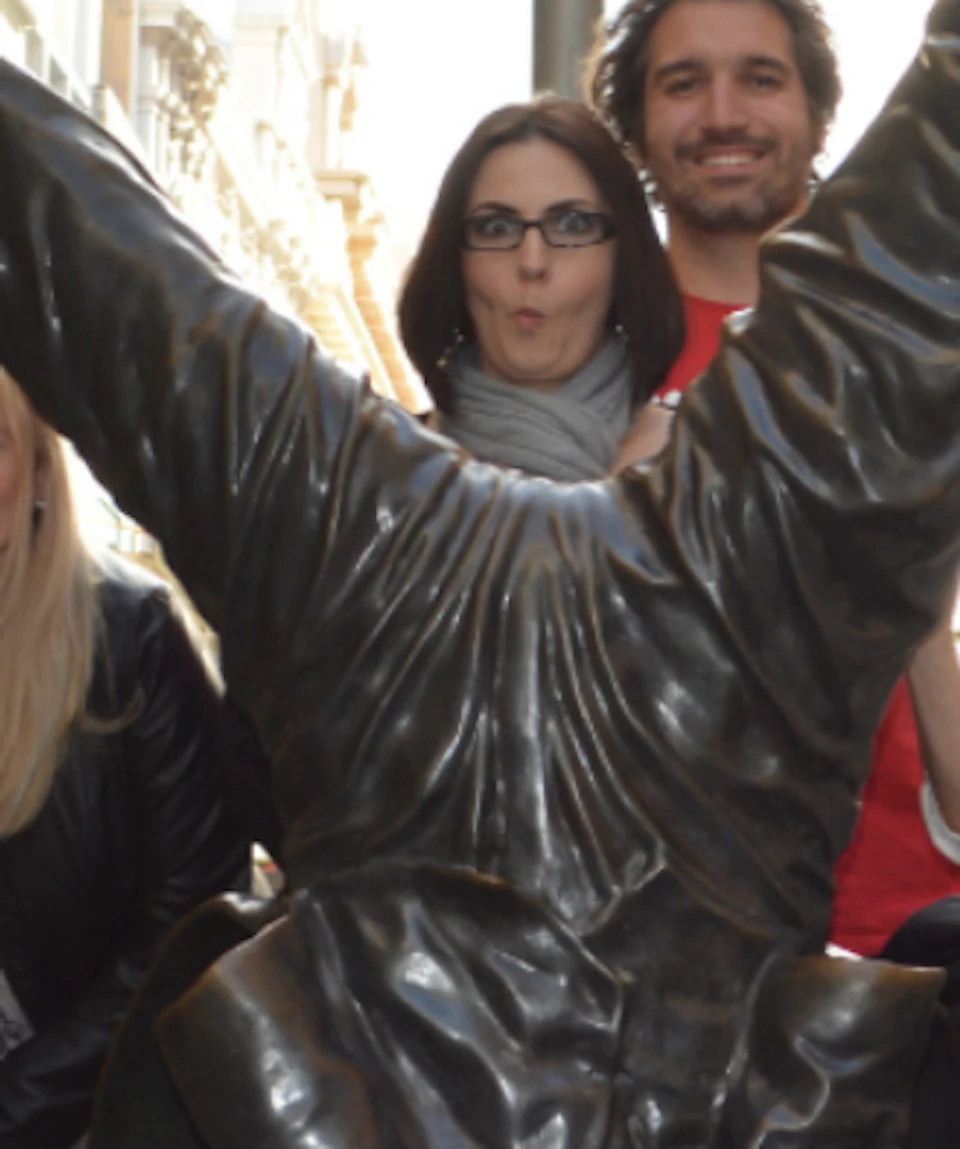Group of people posing around an upside-down statue on a city street, with buildings and shop signs in the background.