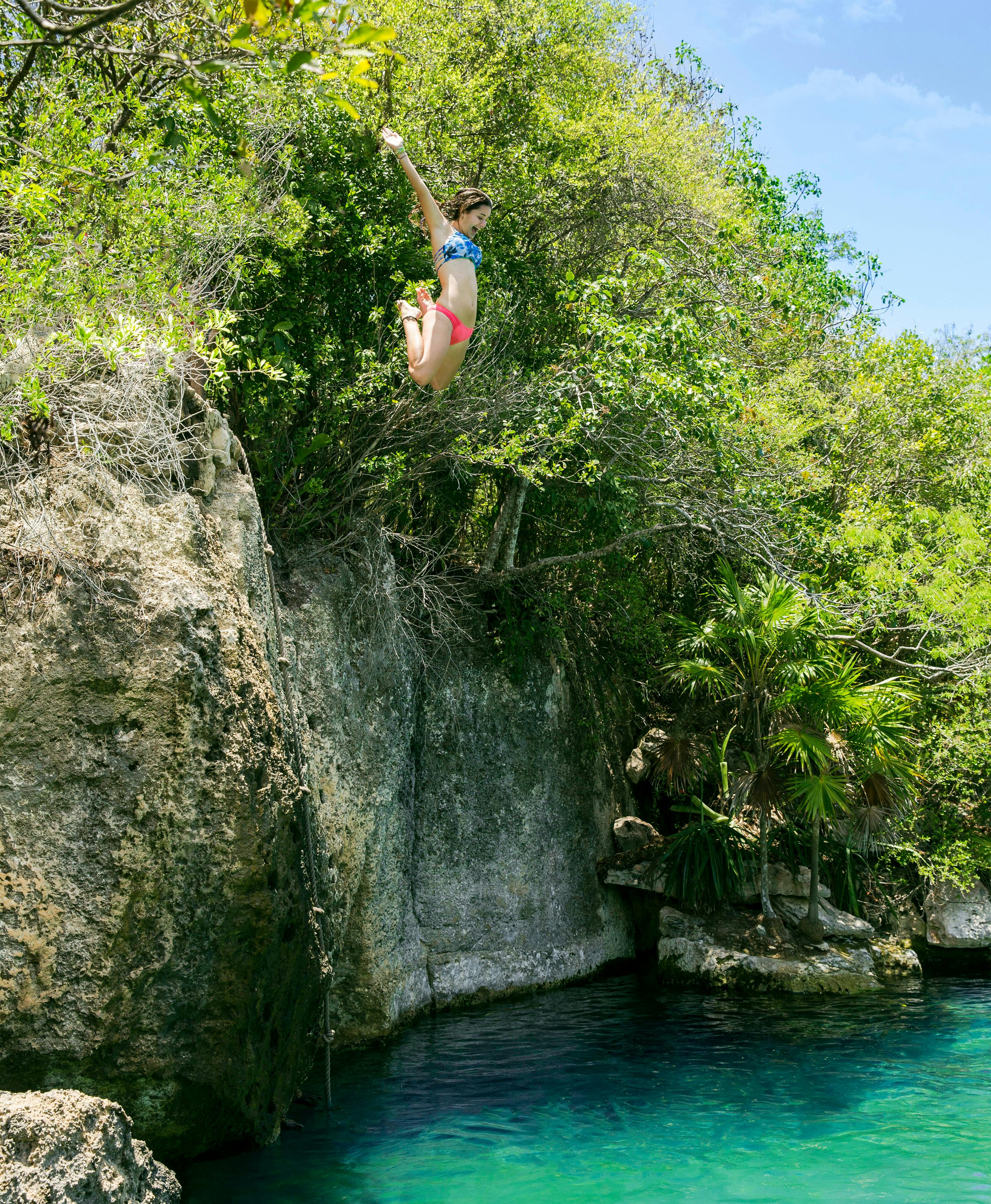Une personne en maillot de bain saute d'une falaise rocheuse dans une étendue d'eau claire et bleue entourée d'une végétation luxuriante.