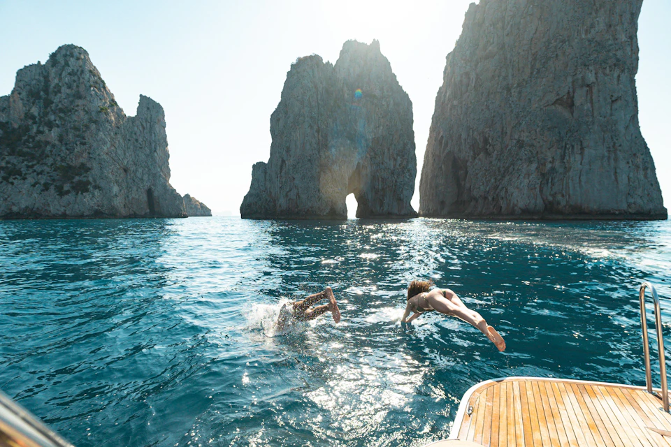 Two people diving off a boat into clear blue water near large rock formations under a sunny sky.
